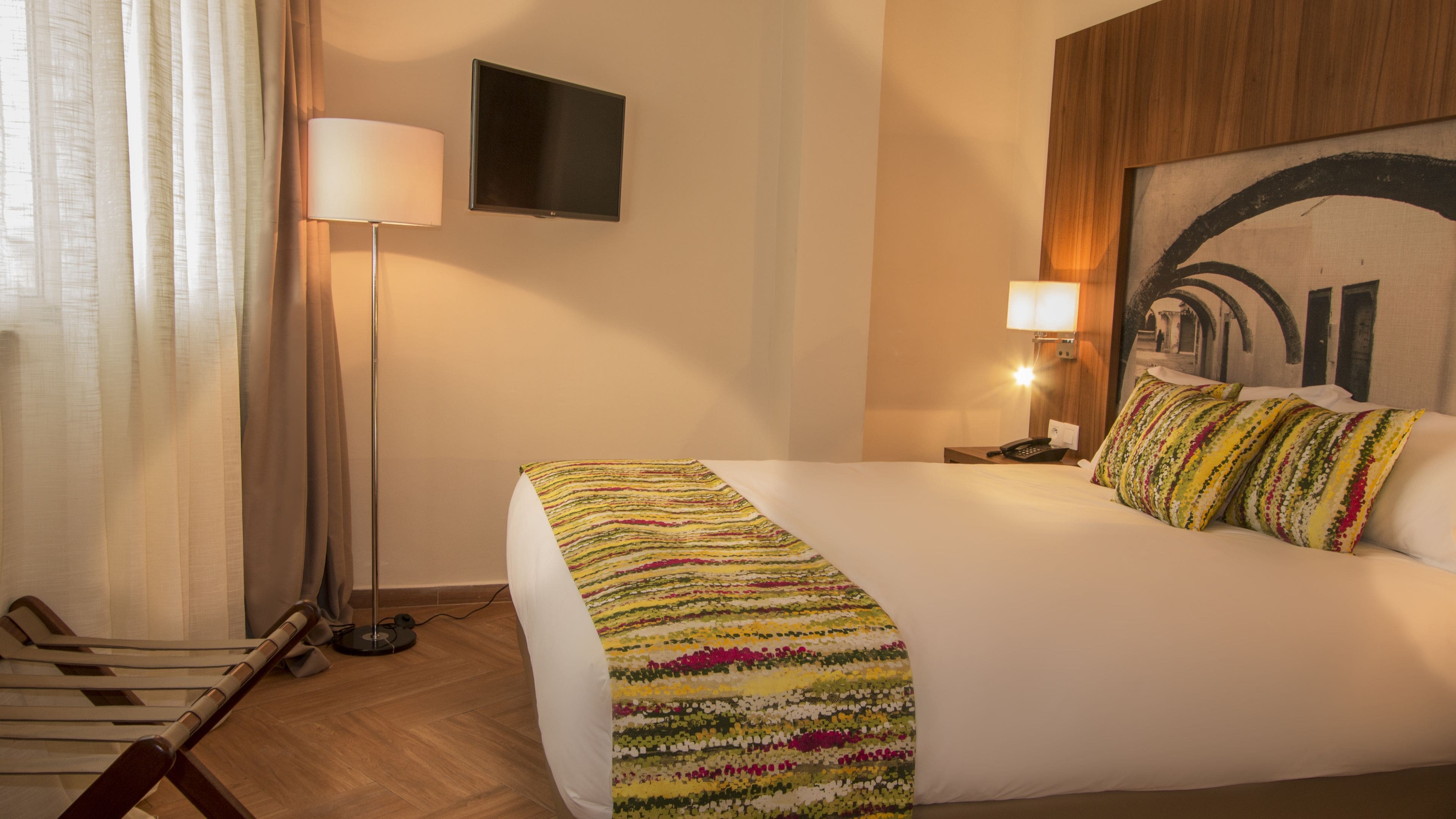 A hotel room with a large bed, a flat-screen TV mounted on the wall, a standing lamp, and a painting on the wall.