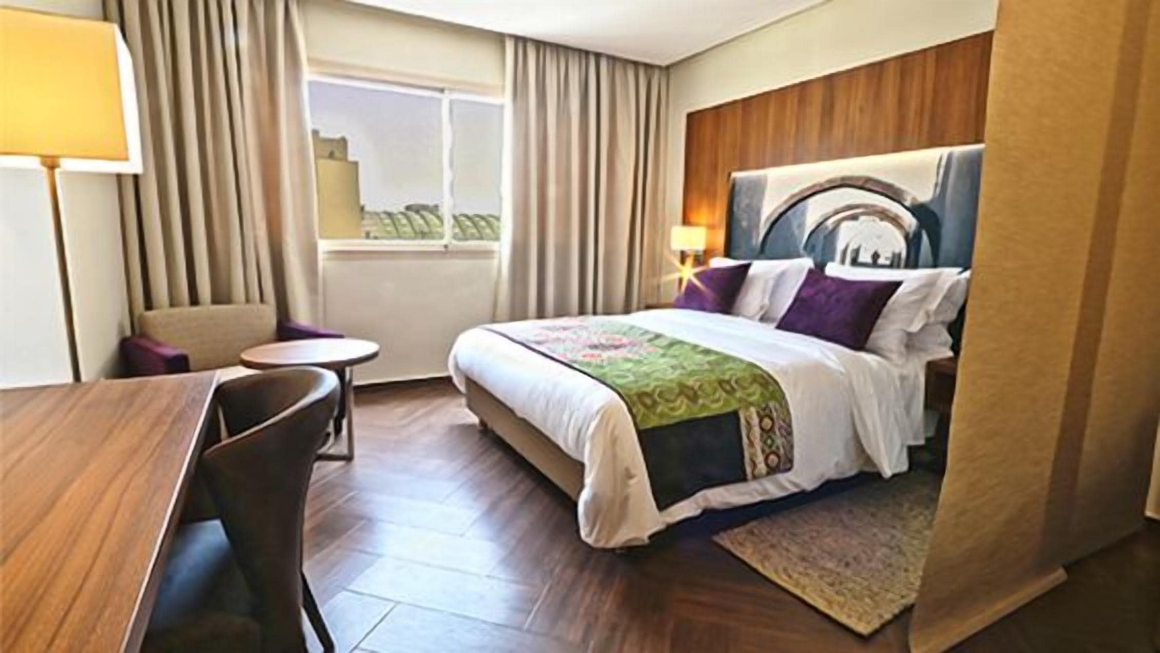 Ein Hotelzimmer mit Bett, Schreibtisch, Stuhl, Nachttisch und einem Fenster mit Vorhängen.