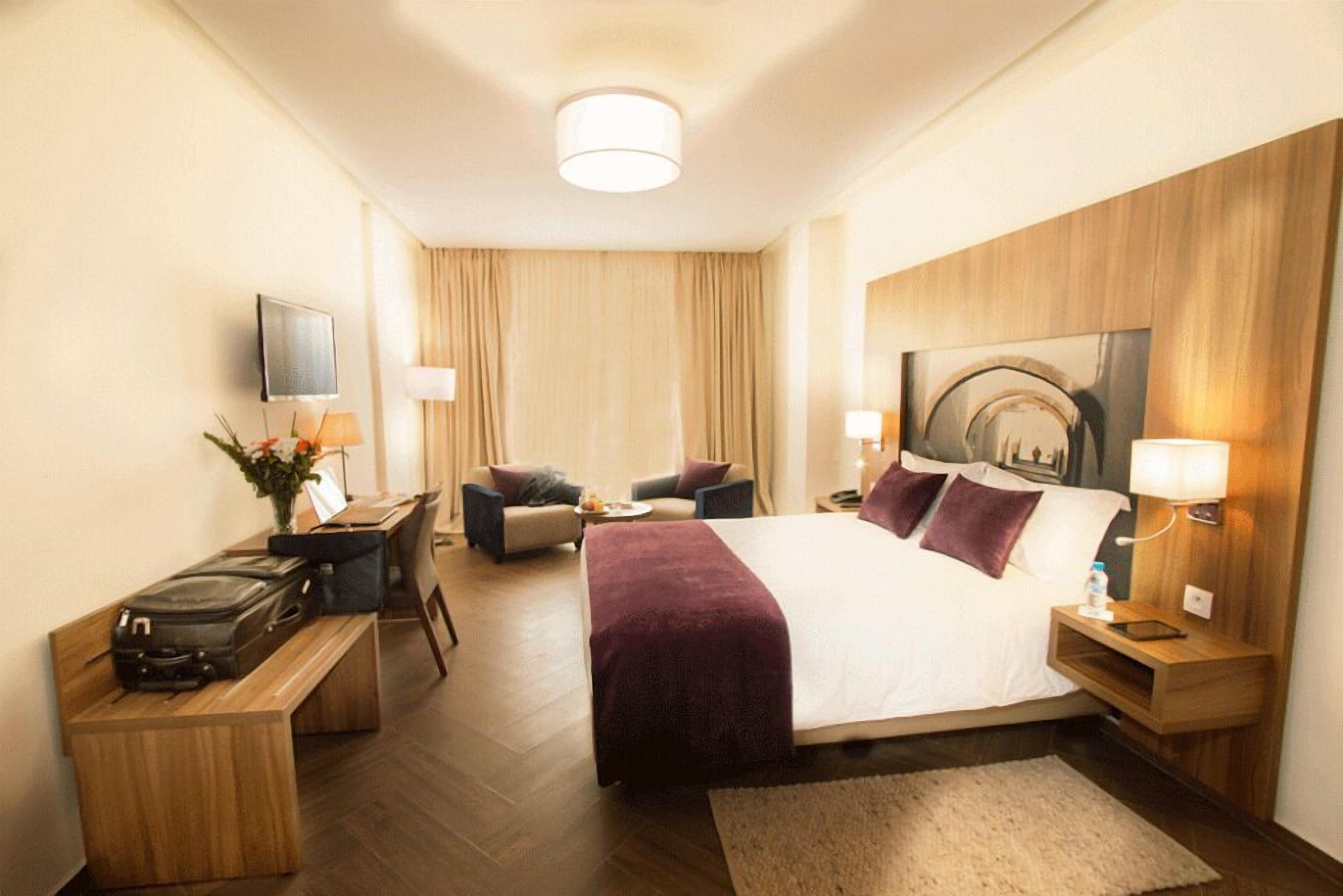 Une chambre d’hôtel avec un grand lit, un bureau avec un ordinateur, une chaise, une télévision et une tête de lit en bois.