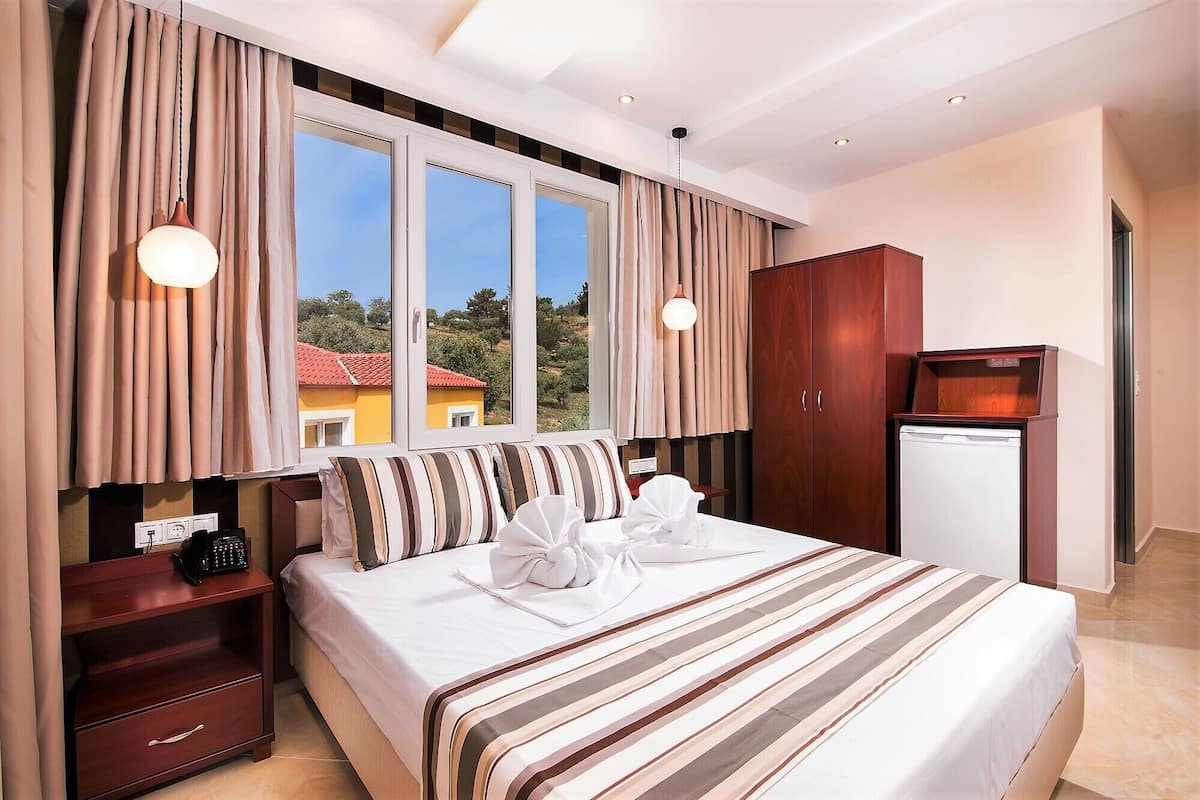 A hotel room with a large bed, two bedside tables, a wardrobe, a desk with a lamp, and a view of the outside.