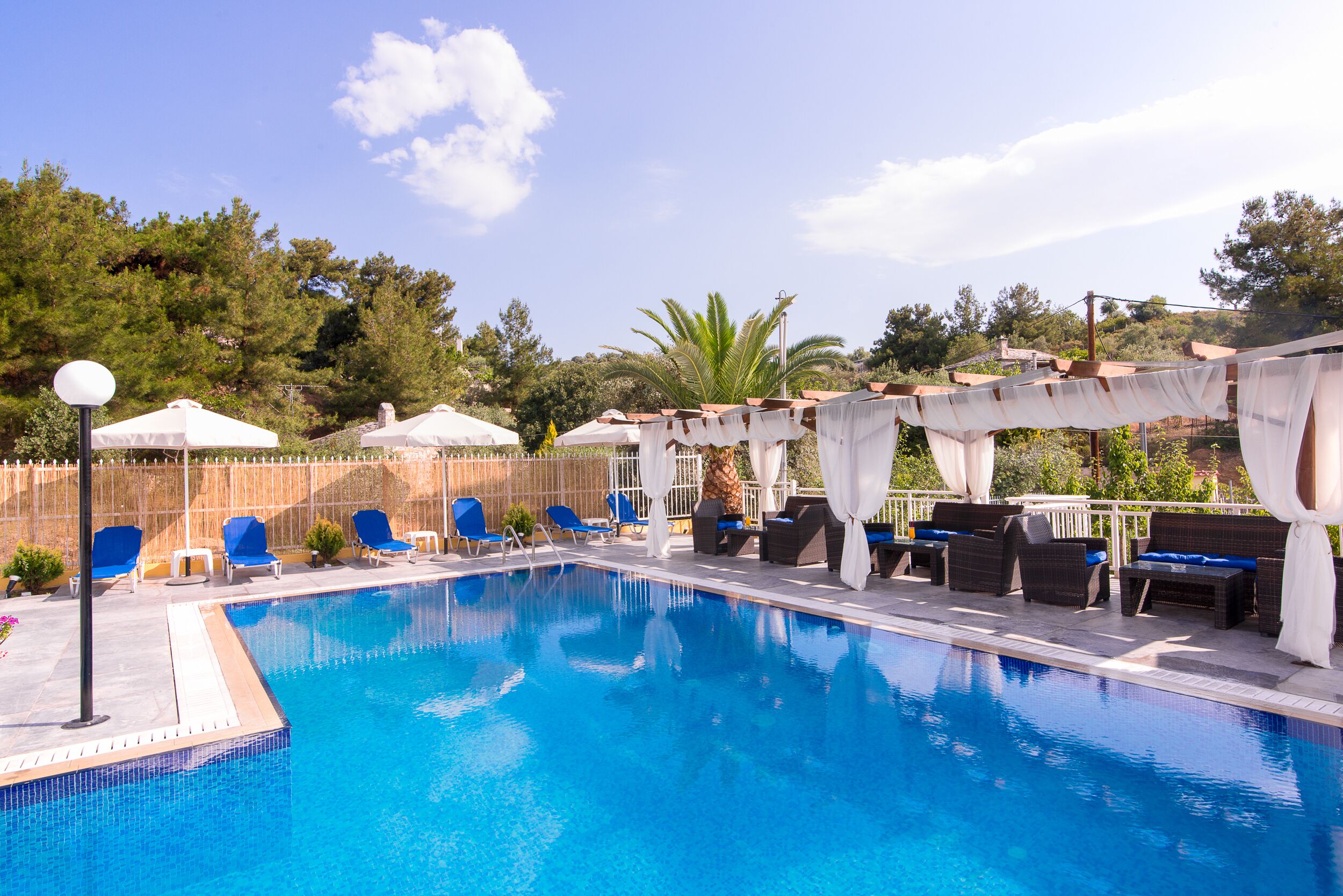 A swimming pool with blue water, surrounded by lounge chairs and umbrellas, set against a backdrop of trees and hills.