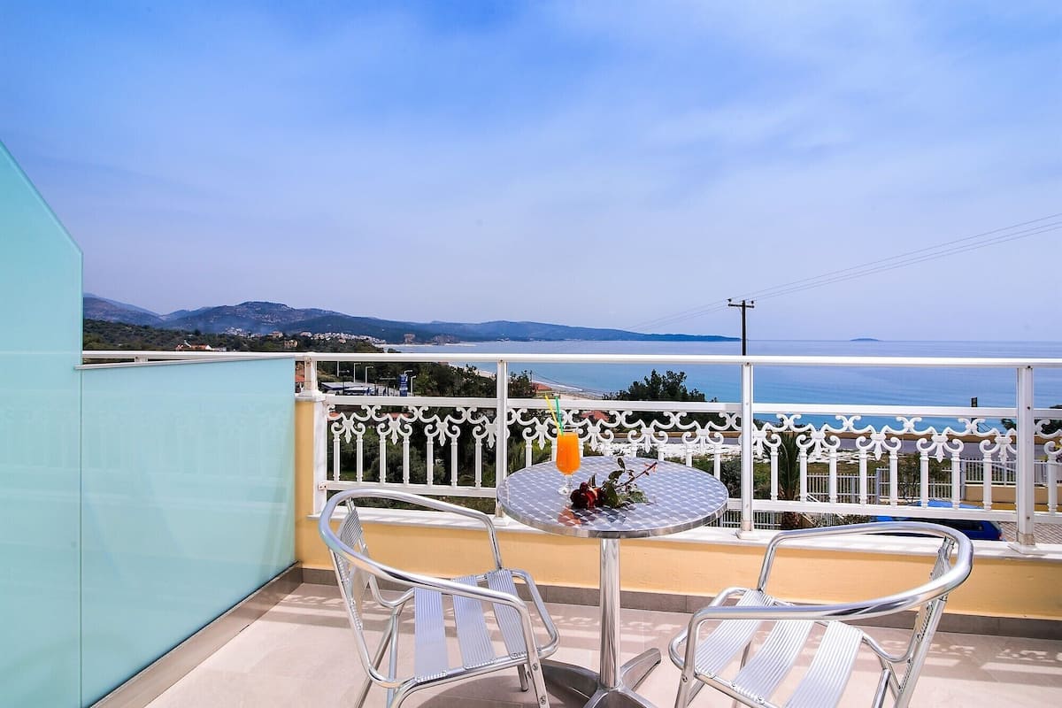 A balcony with two metal chairs and a table, overlooking a coastal view with mountains and a clear sky.