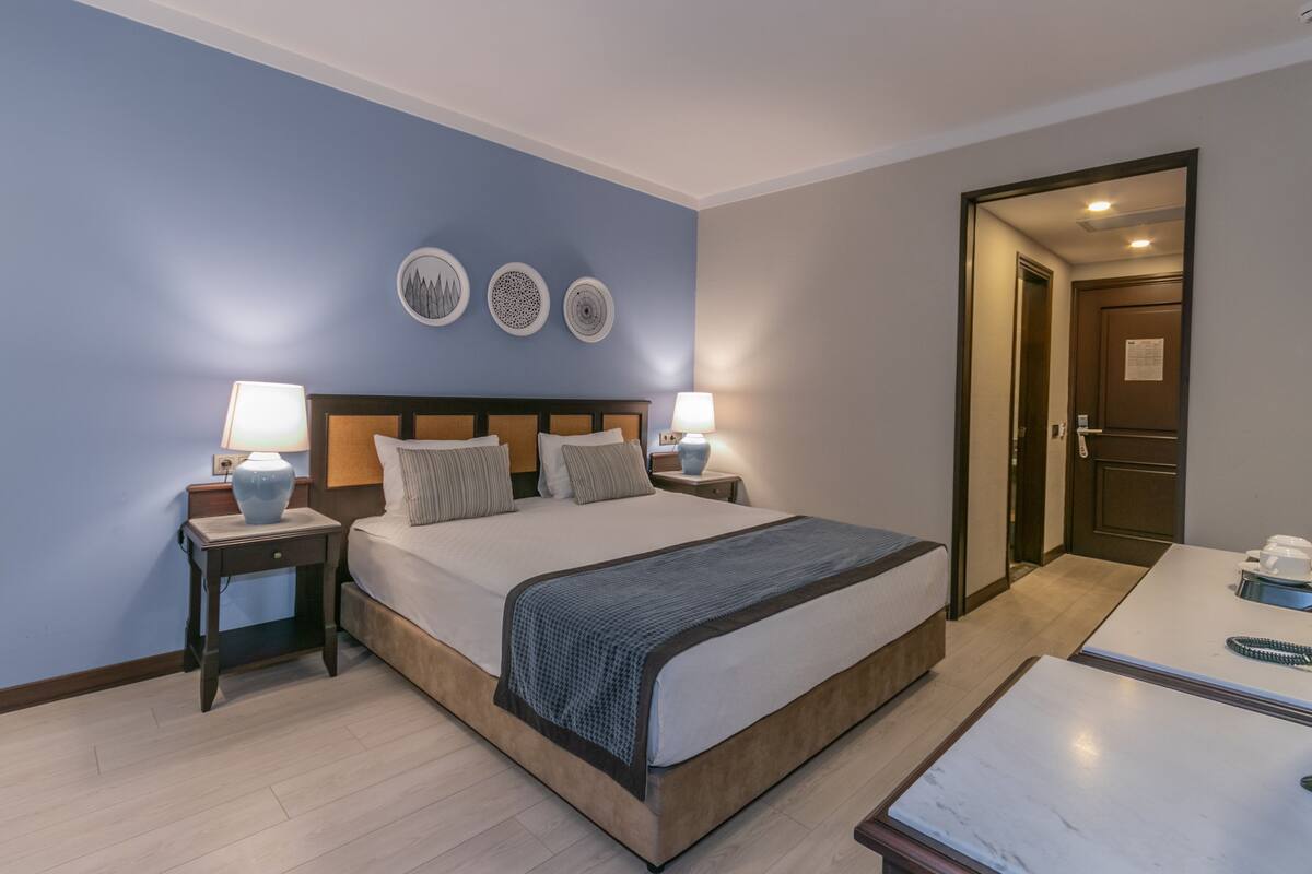 Une chambre d’hôtel avec un grand lit, deux tables de chevet avec des lampes, une petite table avec un vase et une porte donnant sur une autre pièce.