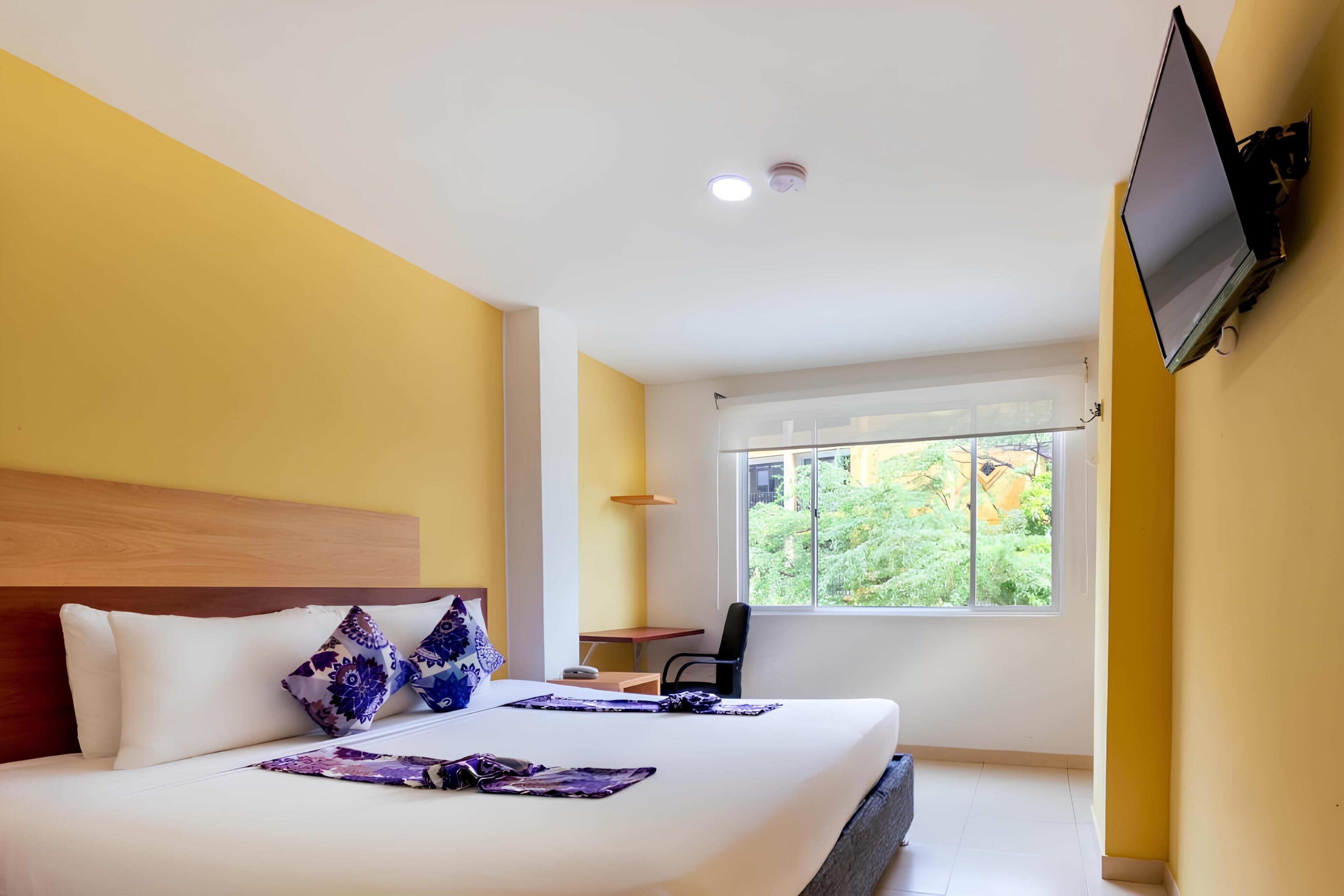 A hotel room with a bed, a TV mounted on the wall, a small desk, and a window with a view of greenery.