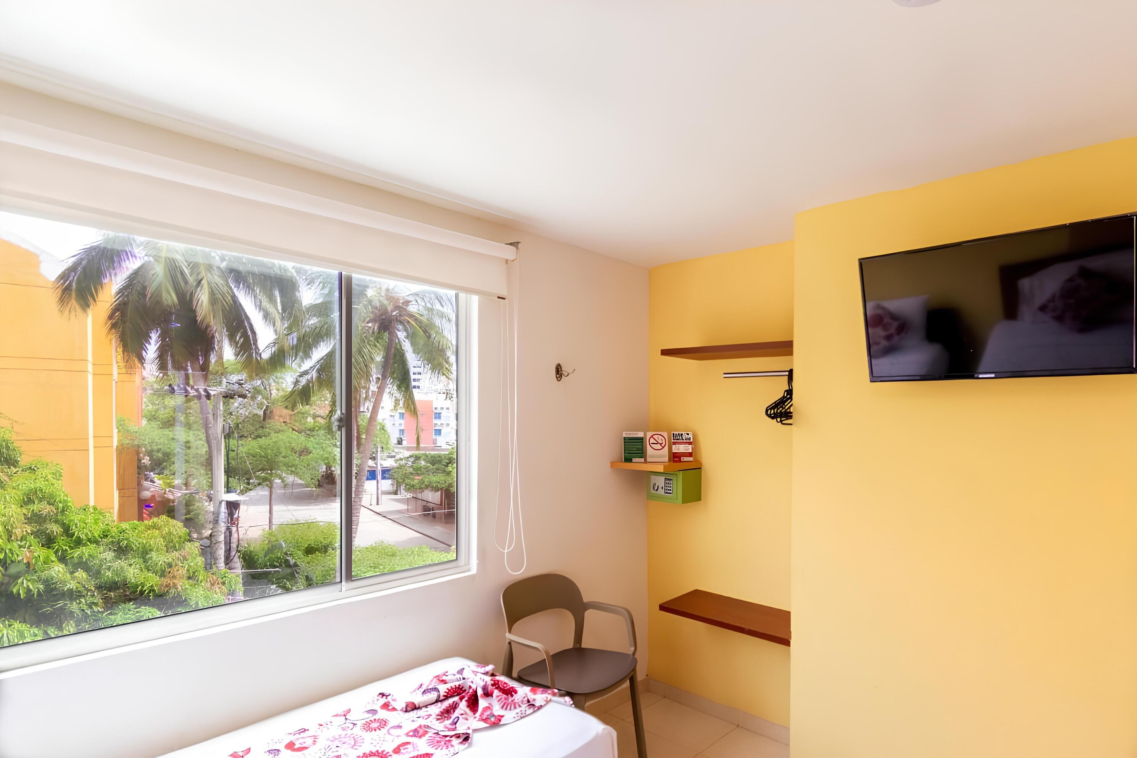 A room with a bed, a chair, a TV, and a window with a view of palm trees and buildings.