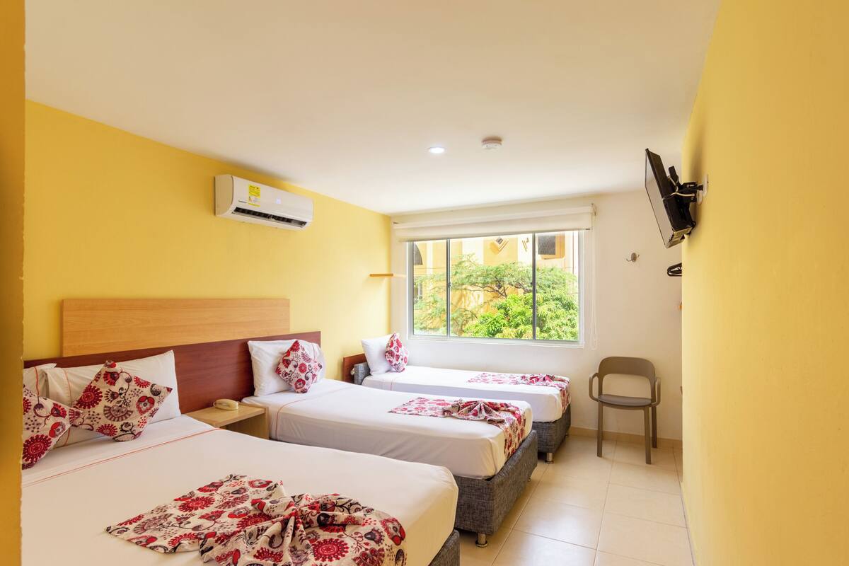Habitación de hotel con tres camas, un televisor montado en la pared, un equipo de aire acondicionado y una ventana con vistas a zonas verdes.