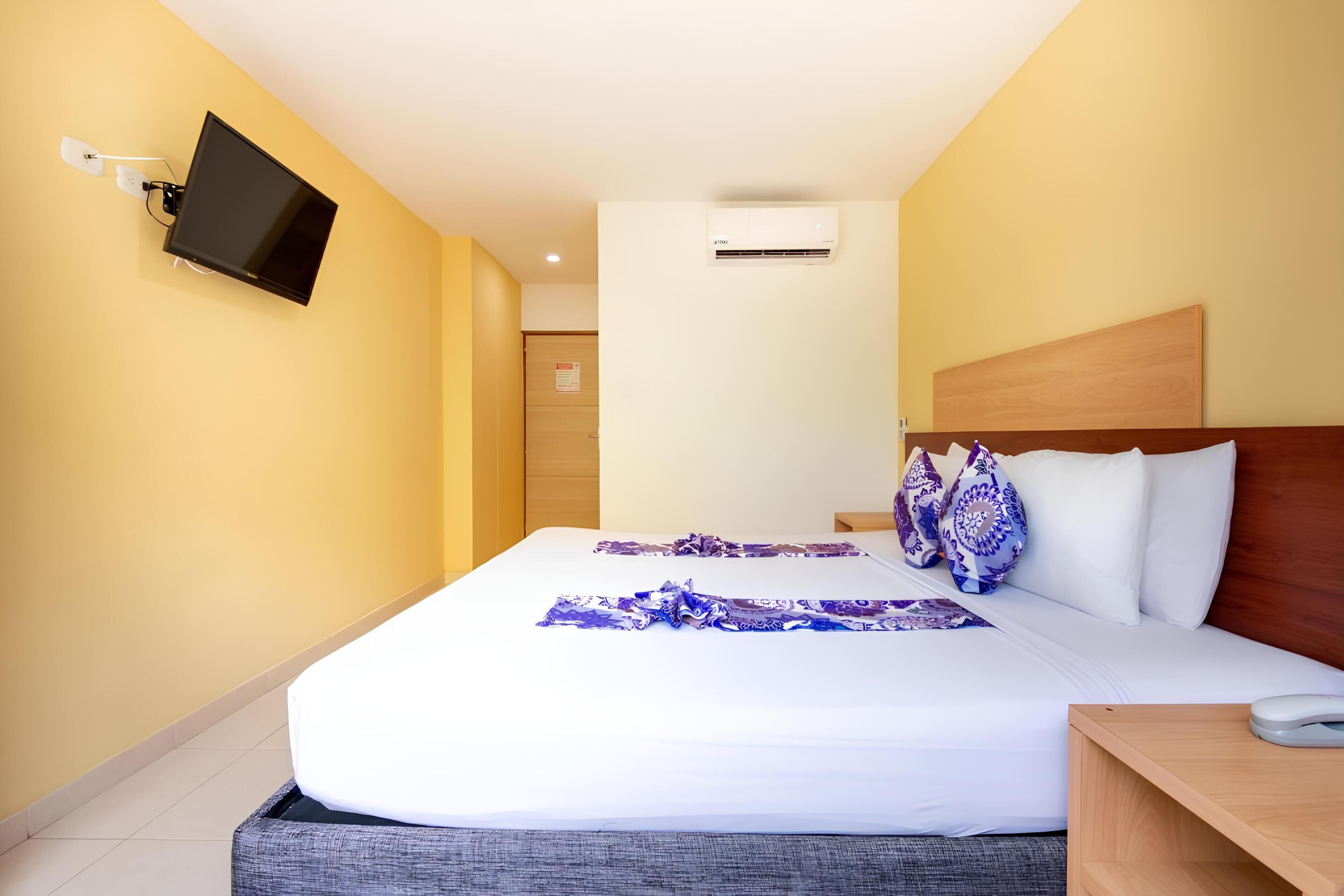 A hotel room with a bed, a mounted TV, an air conditioning unit, and a bedside table.