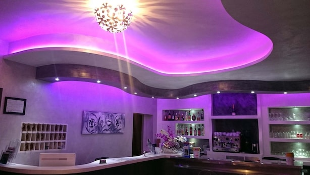 Zona de bar con techo curvo, iluminación morada y una lámpara de araña. Hay estantes con botellas y vasos, y una vitrina de bolsos en la pared.
