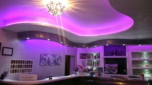 Zona de bar con techo curvo, iluminación morada y una lámpara de araña. Hay estantes con botellas y vasos, y una vitrina de bolsos en la pared.