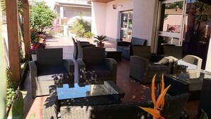Un patio con mobiliario de mimbre y una mesa de vidrio, rodeado de macetas con plantas y vistas a la calle.