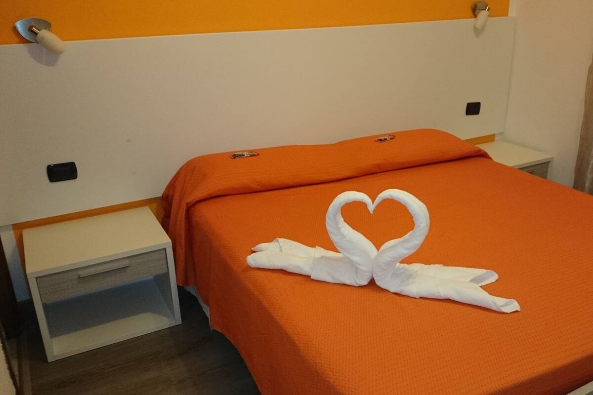 Une chambre d’hôtel avec une couvre-lit orange, une disposition de serviettes en forme de cœur, une petite table de chevet et une commode.