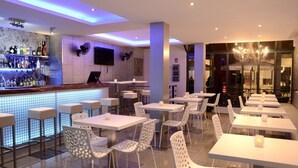 A modern bar area with a counter, shelves of bottles, and a TV screen. There are white tables and chairs arranged for dining.