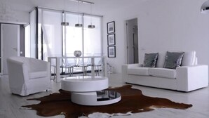 A modern living room with a white sofa, a round coffee table, and a dining area with transparent chairs.