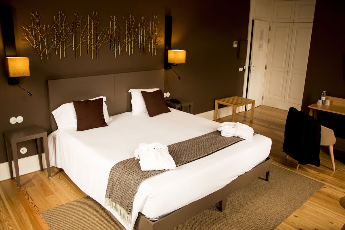 Un lit bien fait, avec du linge de lit blanc et marron, une table de chevet avec une lampe et un luminaire fixé au mur.