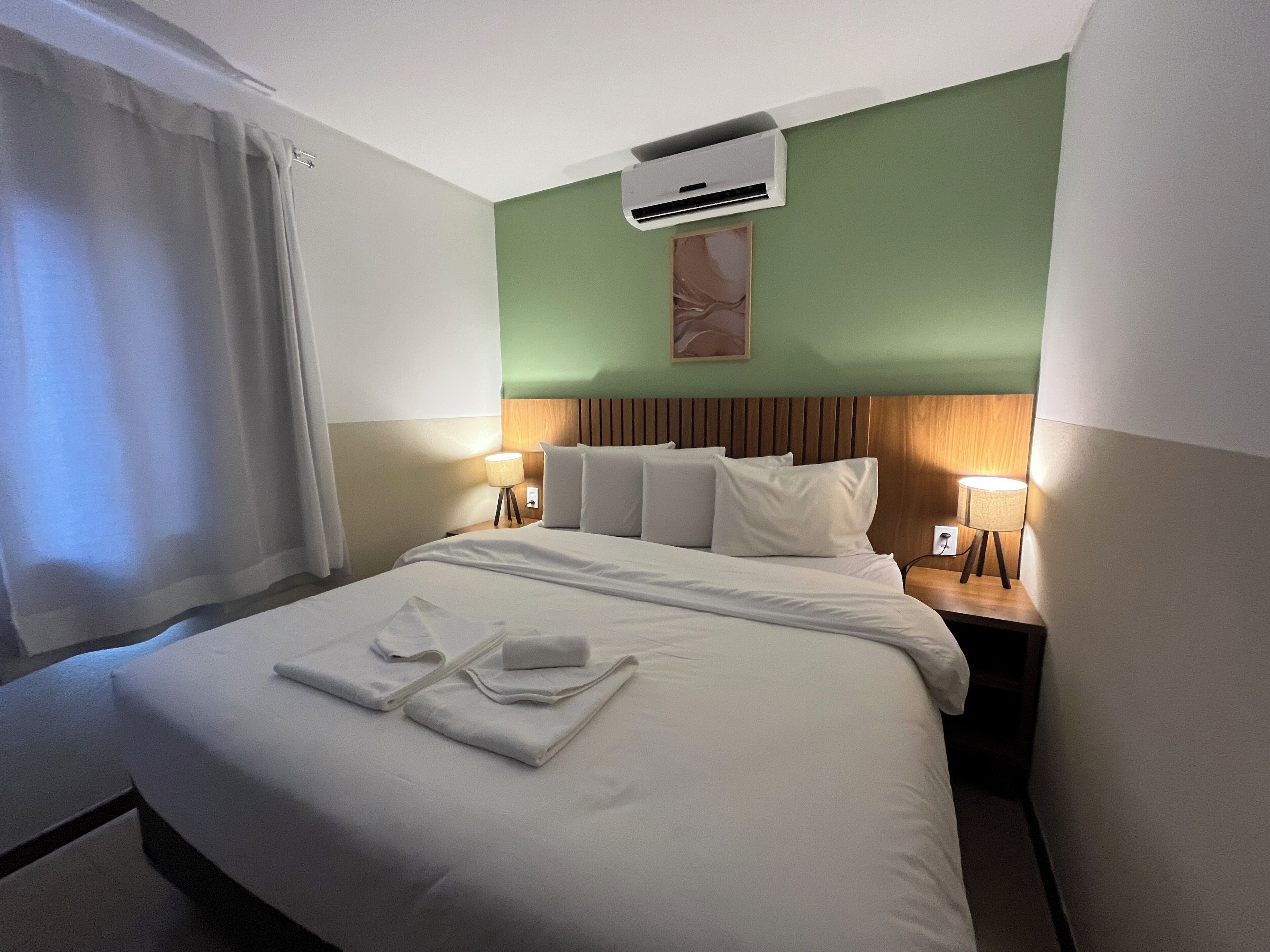 A hotel room with a bed, two bedside lamps, an air conditioning unit, and a window with curtains.