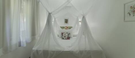 A room with a bed under a white canopy, a small table with a floral tablecloth, and a framed picture on the wall.