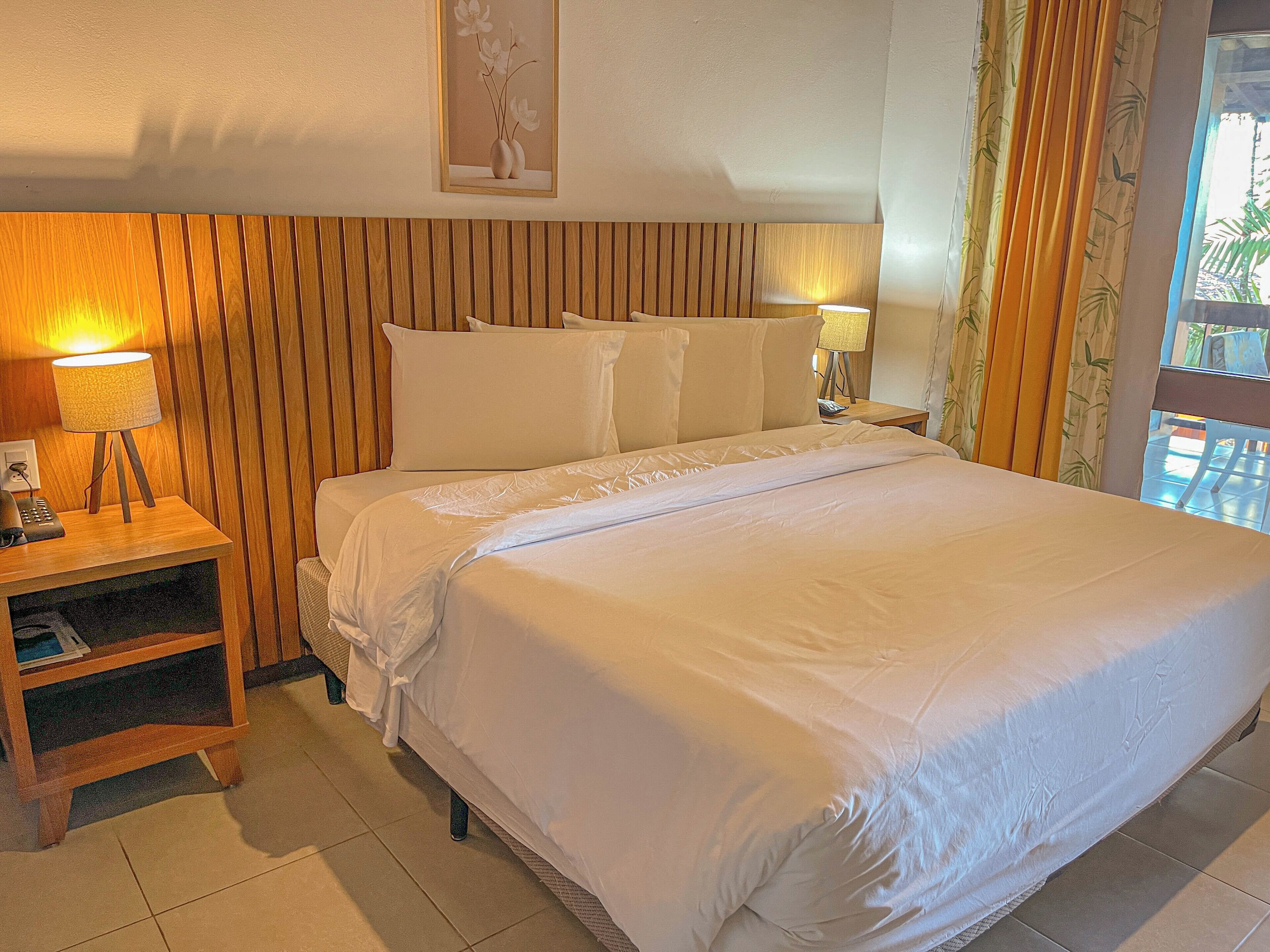 A hotel room with a wooden headboard, a large bed with white linens, a bedside table with a lamp, and a small shelf.