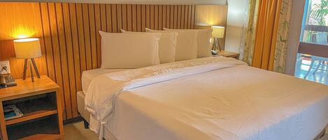 A hotel room with a wooden headboard, a large bed with white linens, a bedside table with a lamp, and a small shelf.