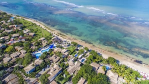 An aerial view of a coastal resort with clear waters, a sandy beach, and a pool area surrounded by bungalow-style accommodations. - Mar Paraíso Hotel (Porto Seguro)