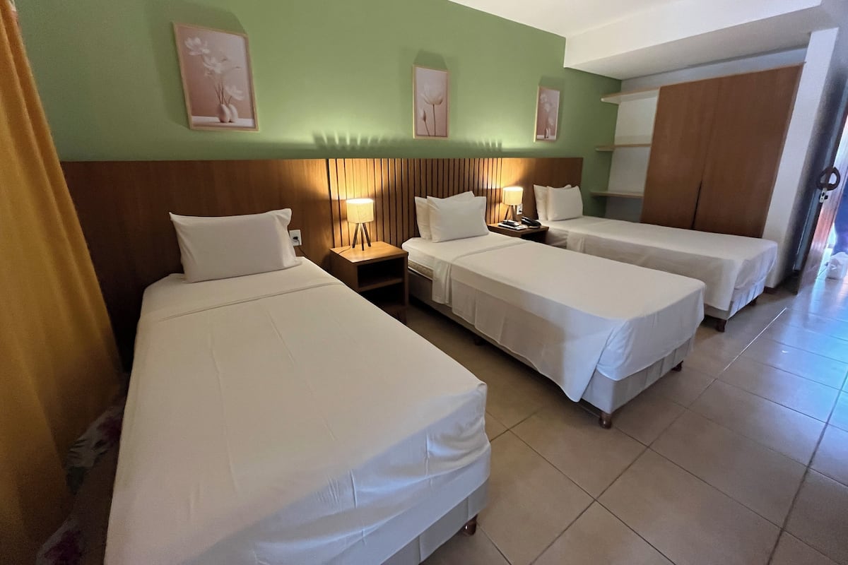A hotel room with three single beds, a wooden headboard, a nightstand with a lamp, and a wardrobe.