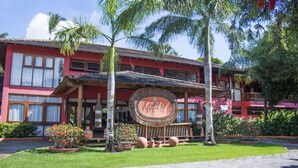 A two-story building with a red facade, a large circular sign with 'Mar Paraiso' written on it, and a well-maintained garden with palm trees and potted plants. - Mar Paraíso Hotel (Porto Seguro)
