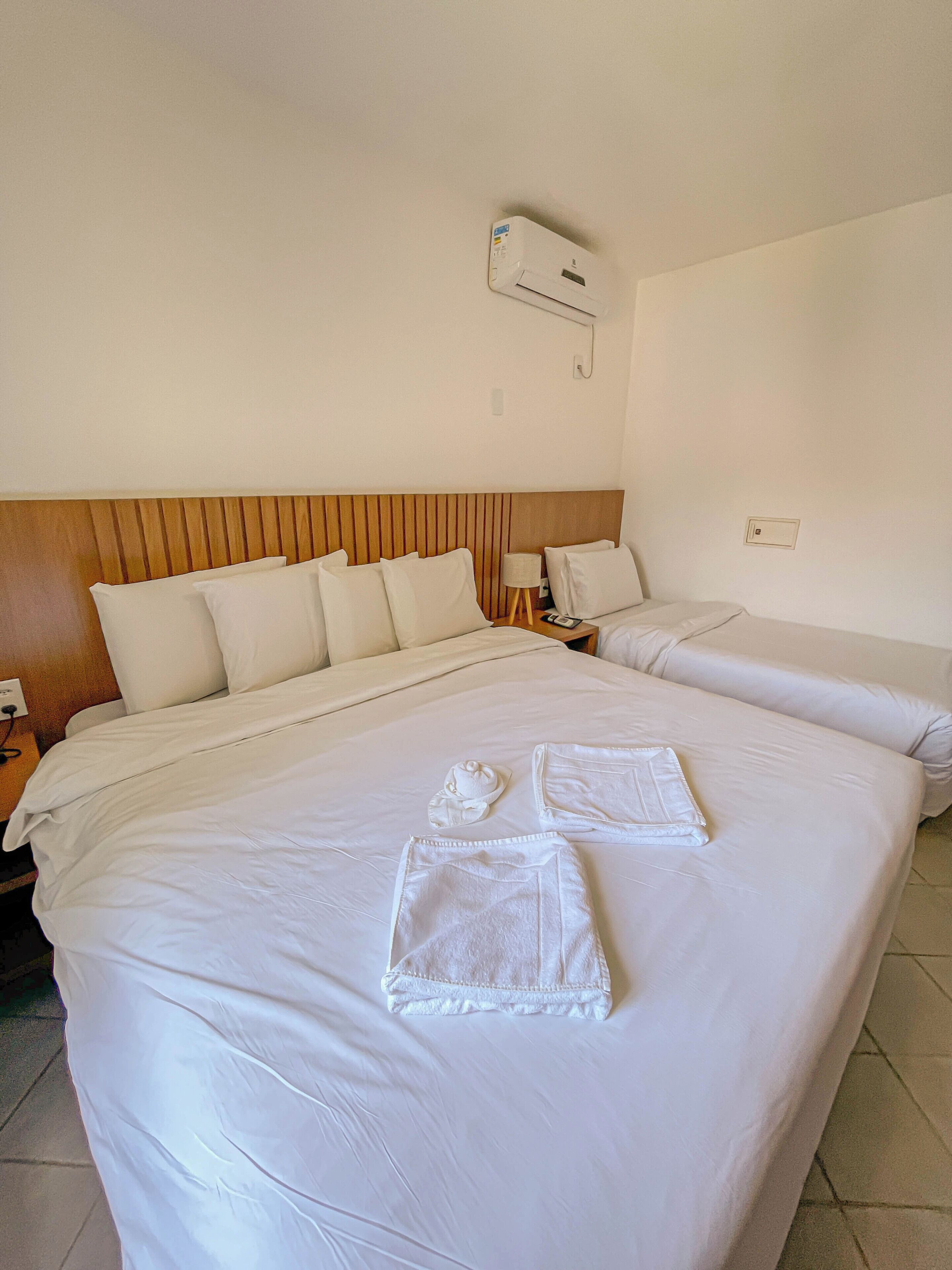 A hotel room with a large bed, a smaller bed, a wooden headboard, a nightstand, a lamp, and an air conditioning unit.