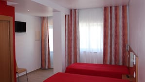 A hotel room with two red beds, a TV mounted on the wall, and a window with striped curtains. - Alojamento Baleal à Vista (Peniche)