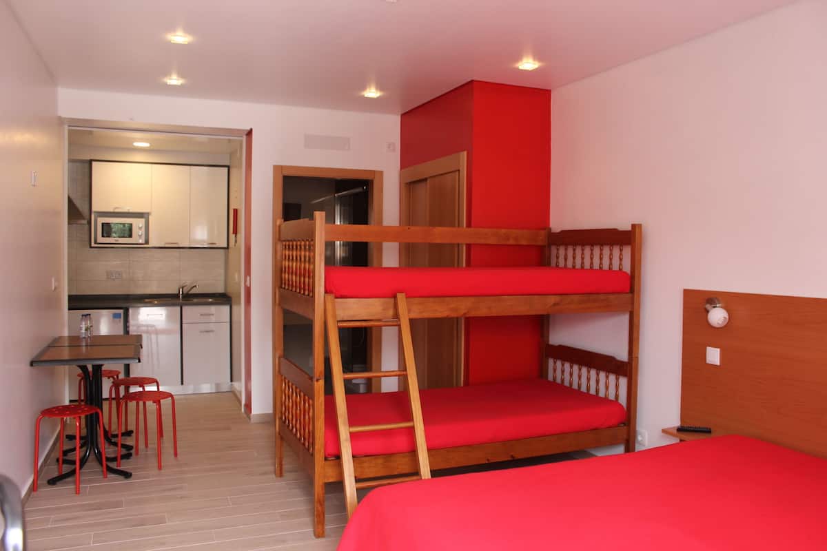 Een stapelbed met rode beddengoed, een houten tafel met stoelen, en een kitchenette op de achtergrond.