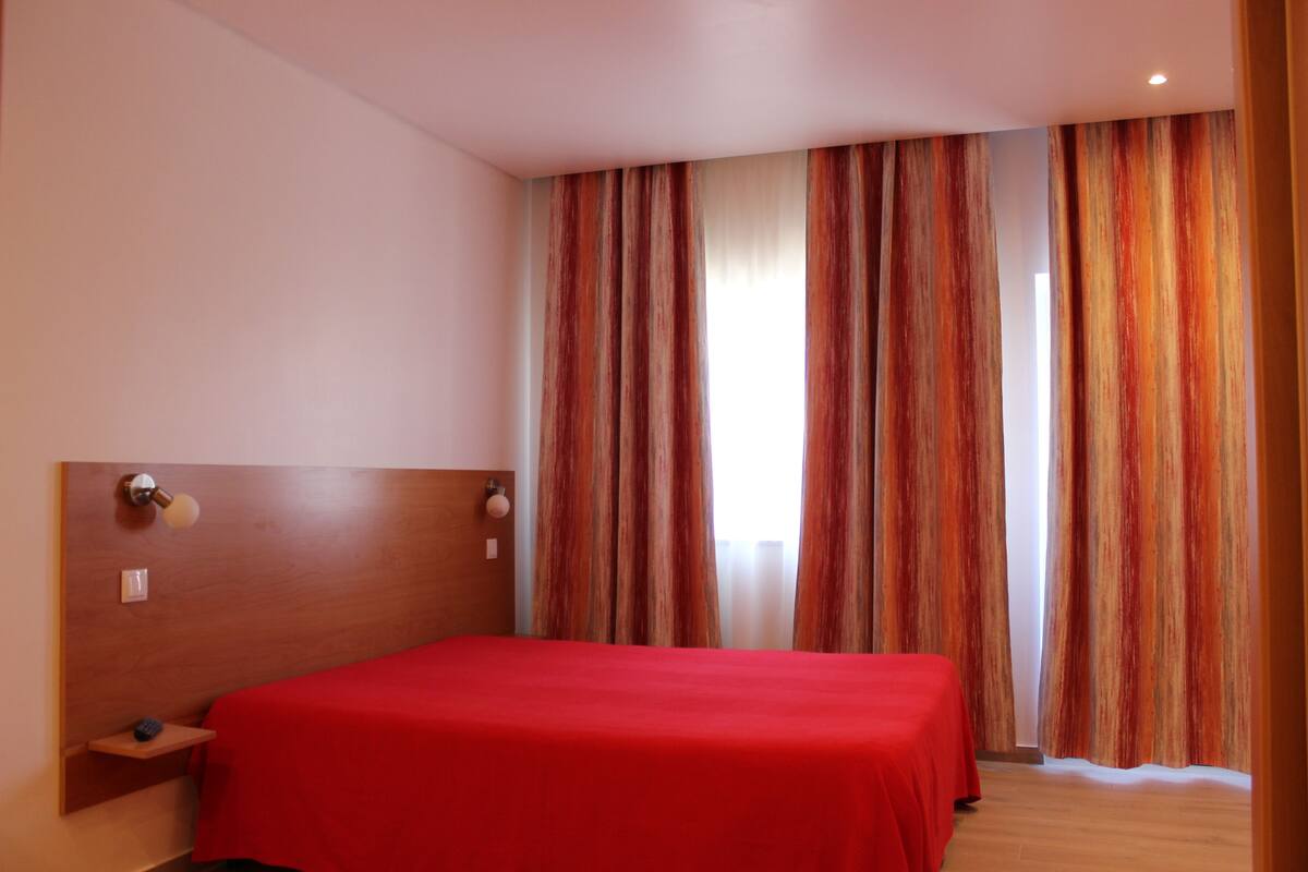 Een hotelkamer met een houten hoofdbord, een rode sprei en een raam met gestreepte gordijnen.