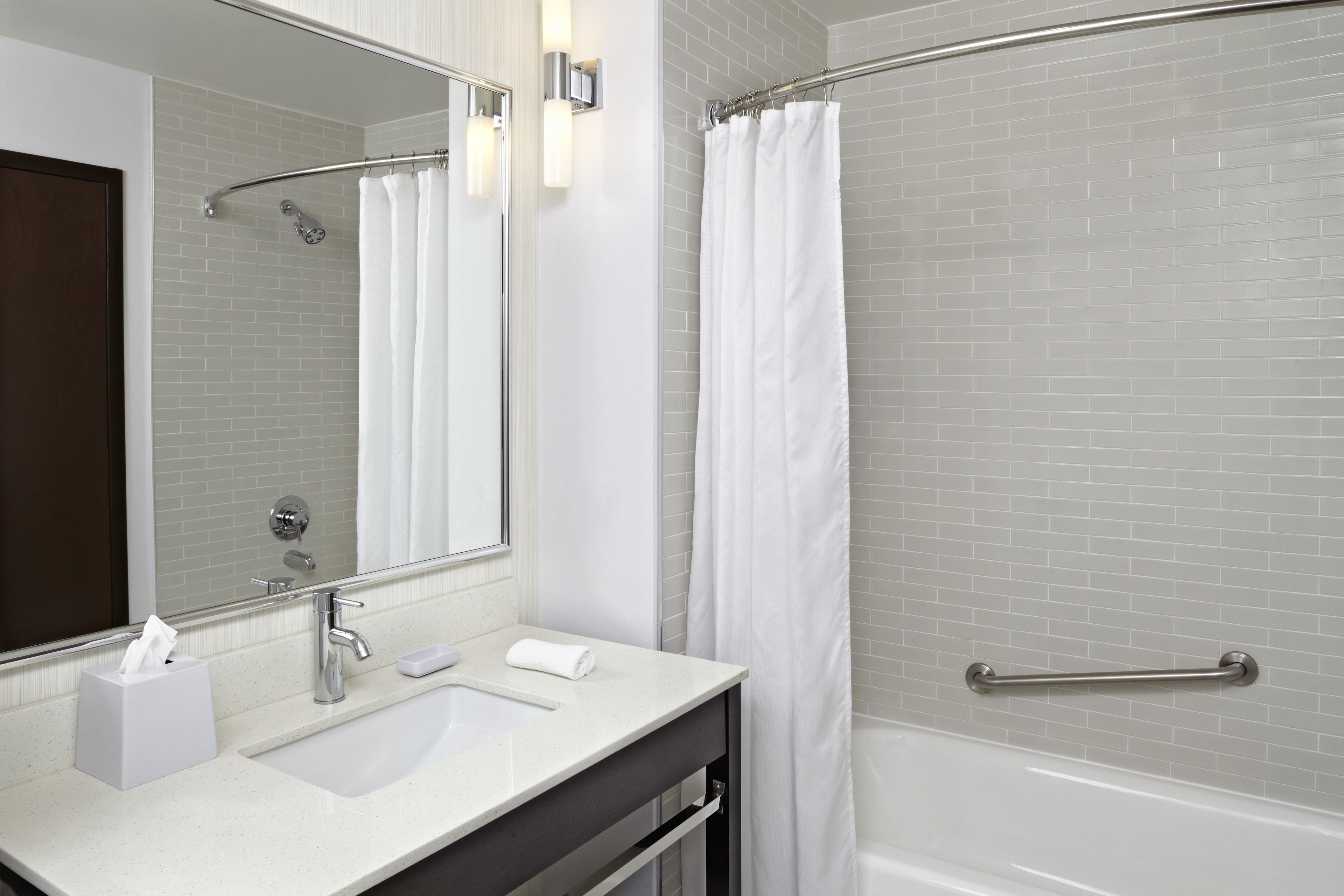 A modern bathroom with a white sink, a large mirror, a shower with a glass door, and a handrail.