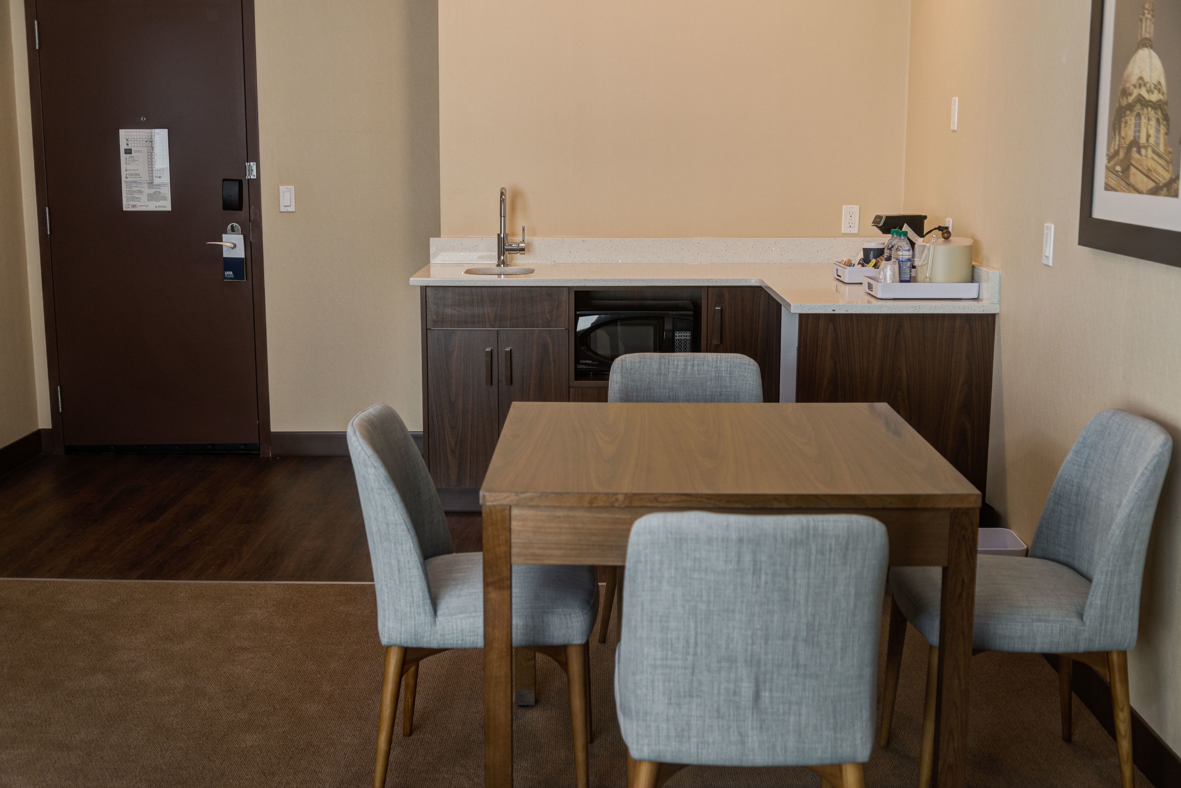 Una cocina compacta con una mesa de madera y cuatro sillas grises, un microondas y un lavamanos.