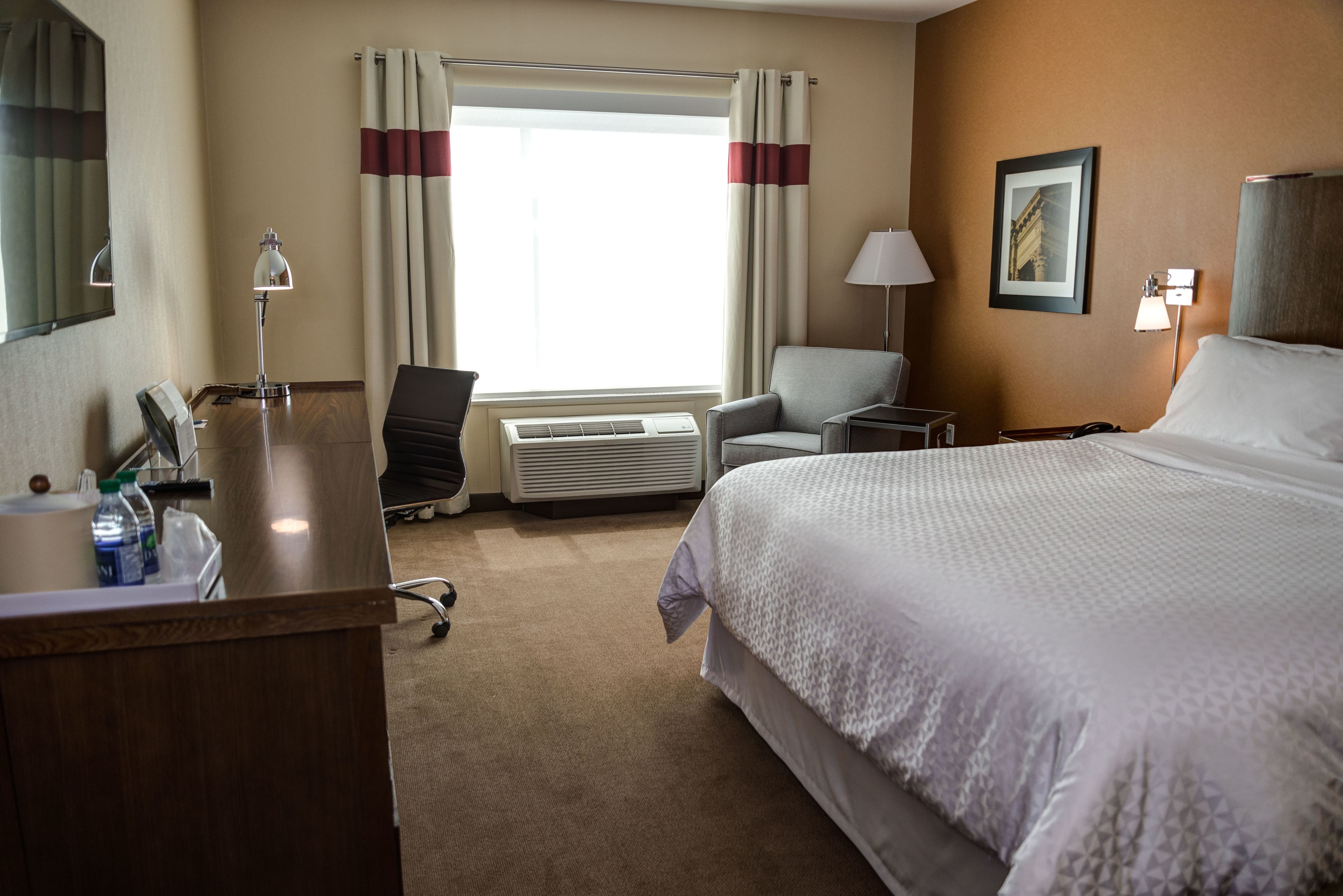A hotel room with a large bed, a desk with a lamp, a chair, a window with curtains, and a picture on the wall.