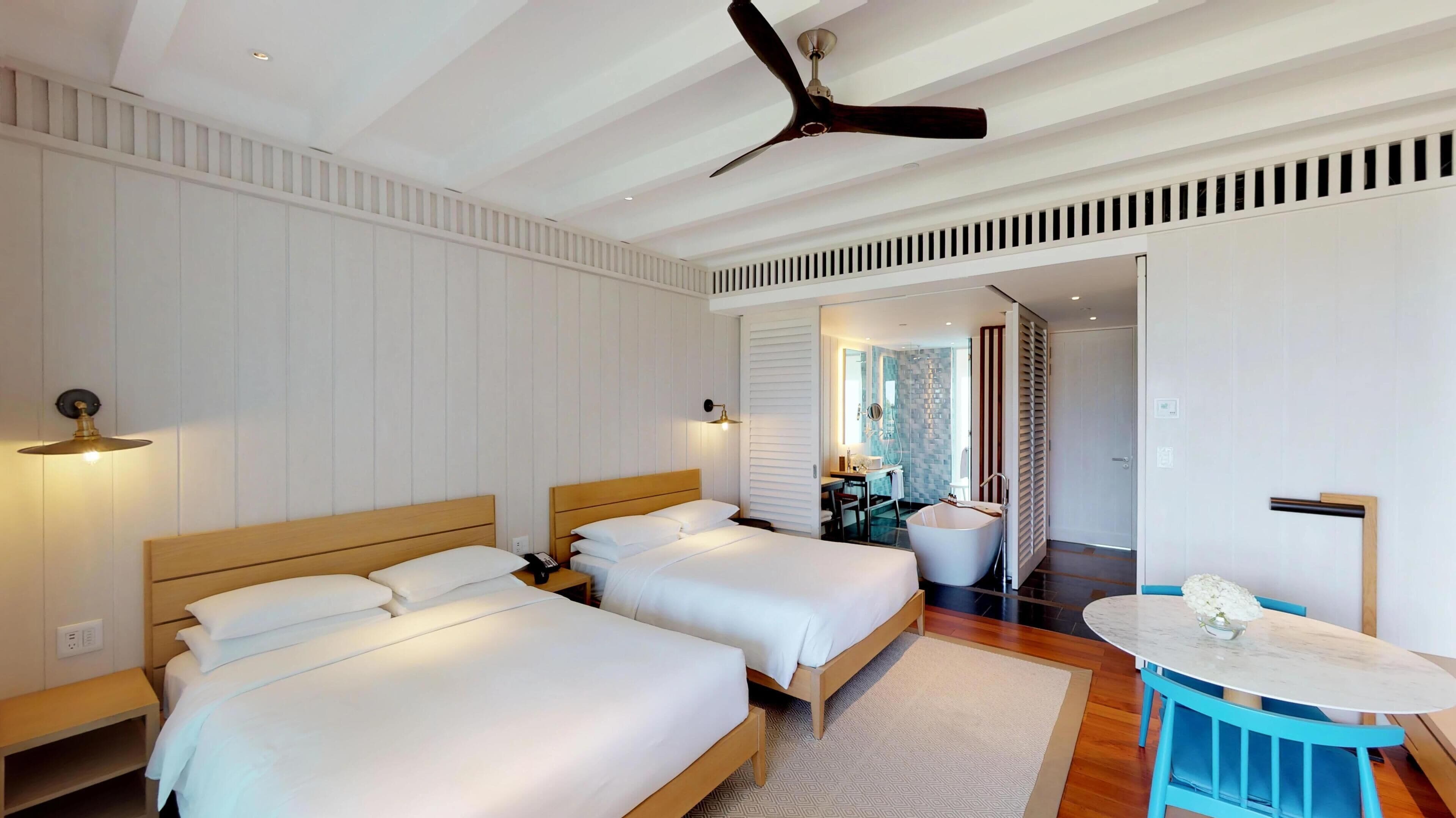 A hotel room with two beds, a ceiling fan, a bathroom with a bathtub, and a small round table with a vase.