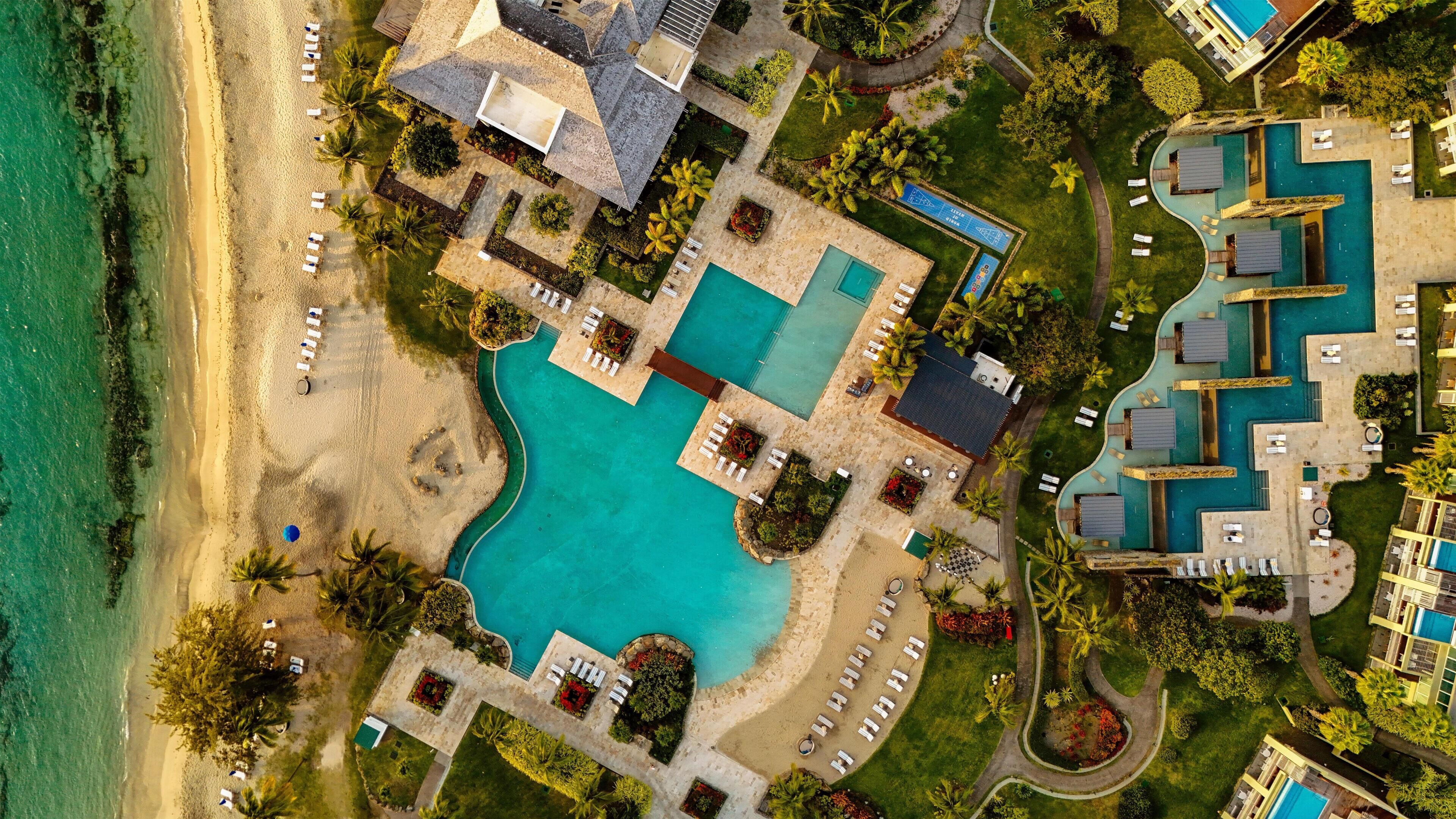 Vista aérea de un complejo turístico con una gran piscina central, varias piscinas más pequeñas y una zona de playa con tumbonas.