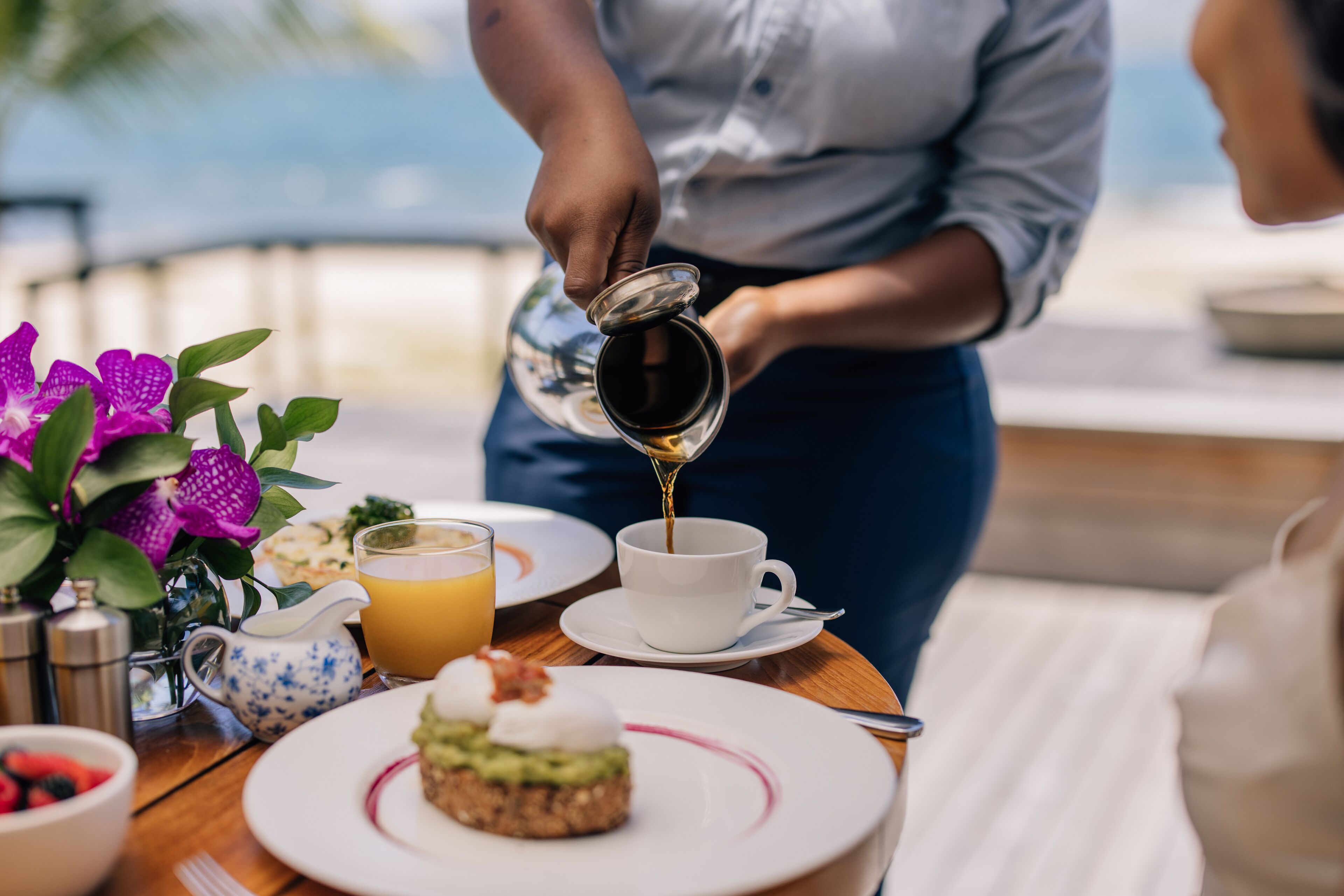A breakfast setting with a person pouring coffee into a cup, a plate with a pastry, and a fruit bowl.