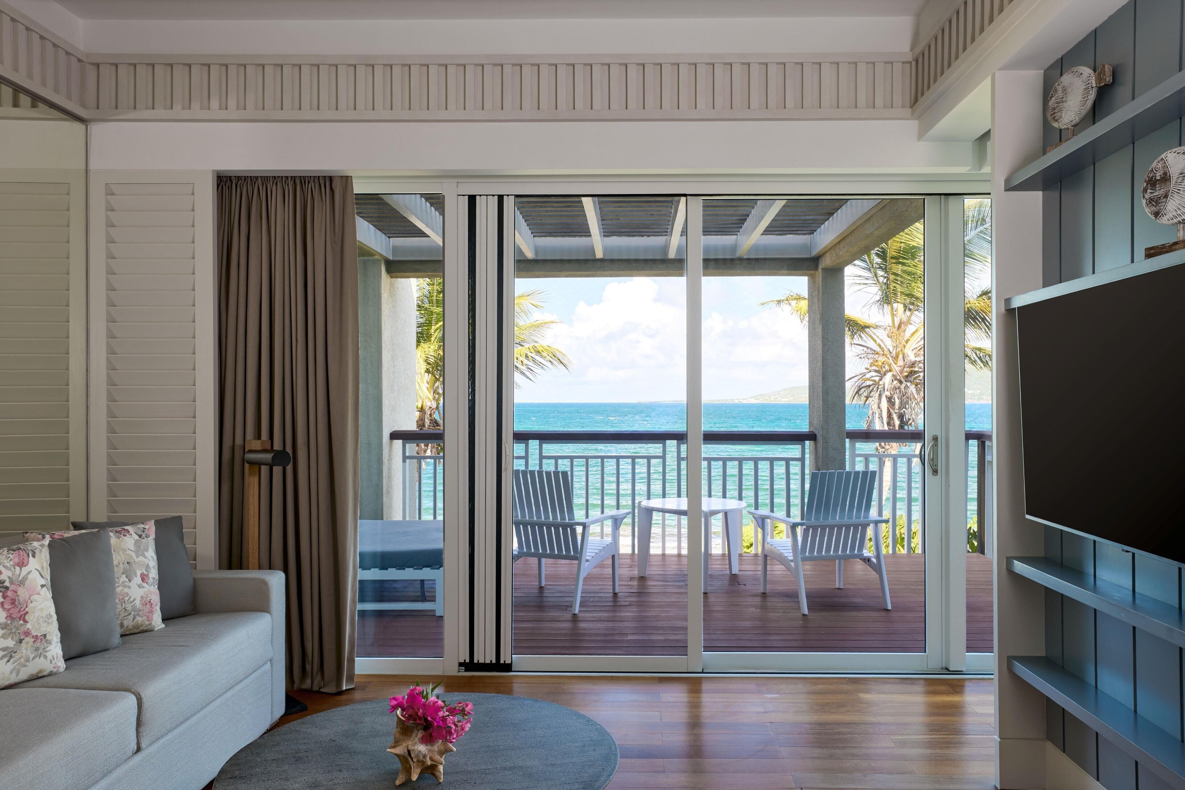 Habitación con sofá, una mesa de centro con flores, televisor y puerta corrediza de vidrio que da a un balcón con sillas y vista al mar.