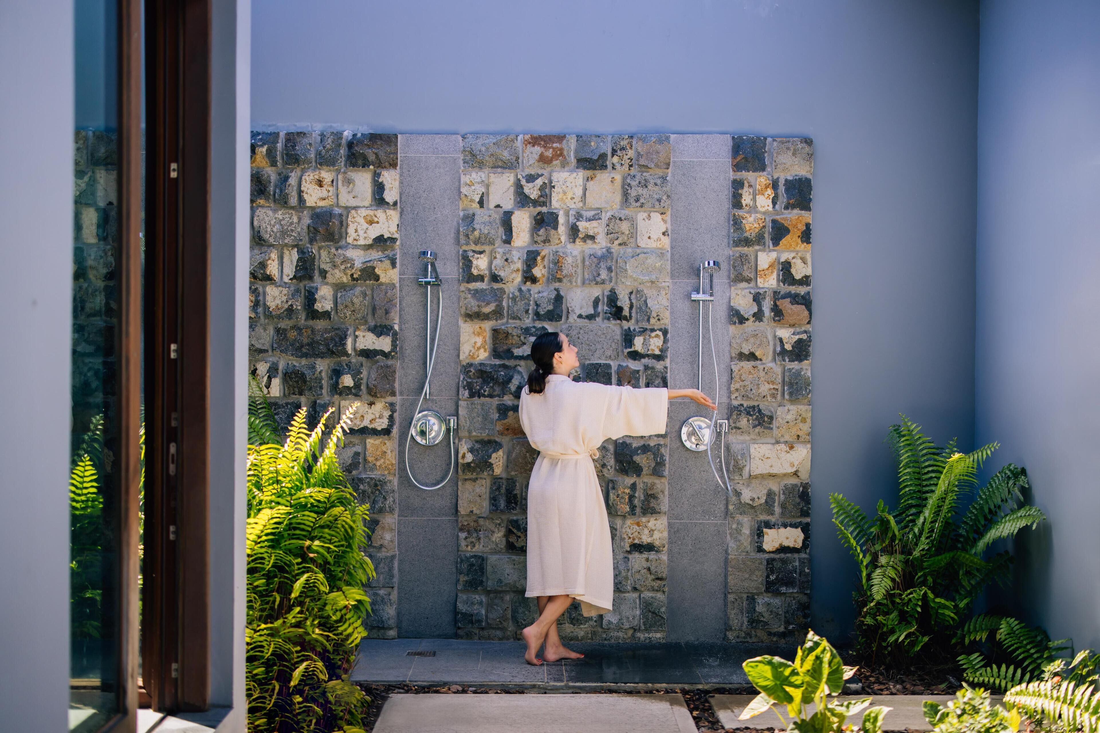 A person in a robe standing in a shower area with stone walls and ferns.