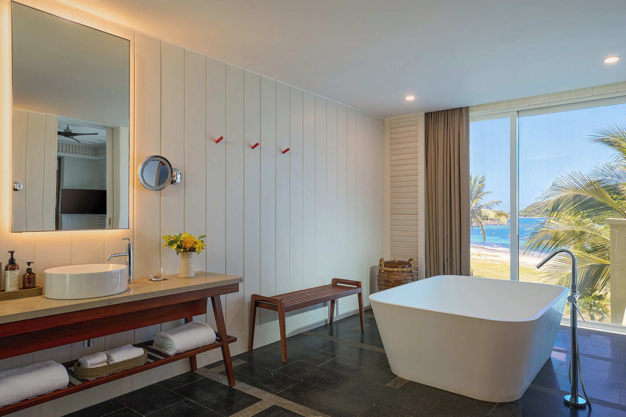 A modern bathroom with a freestanding bathtub, a large mirror, a vanity with a sink, and a view of the beach.