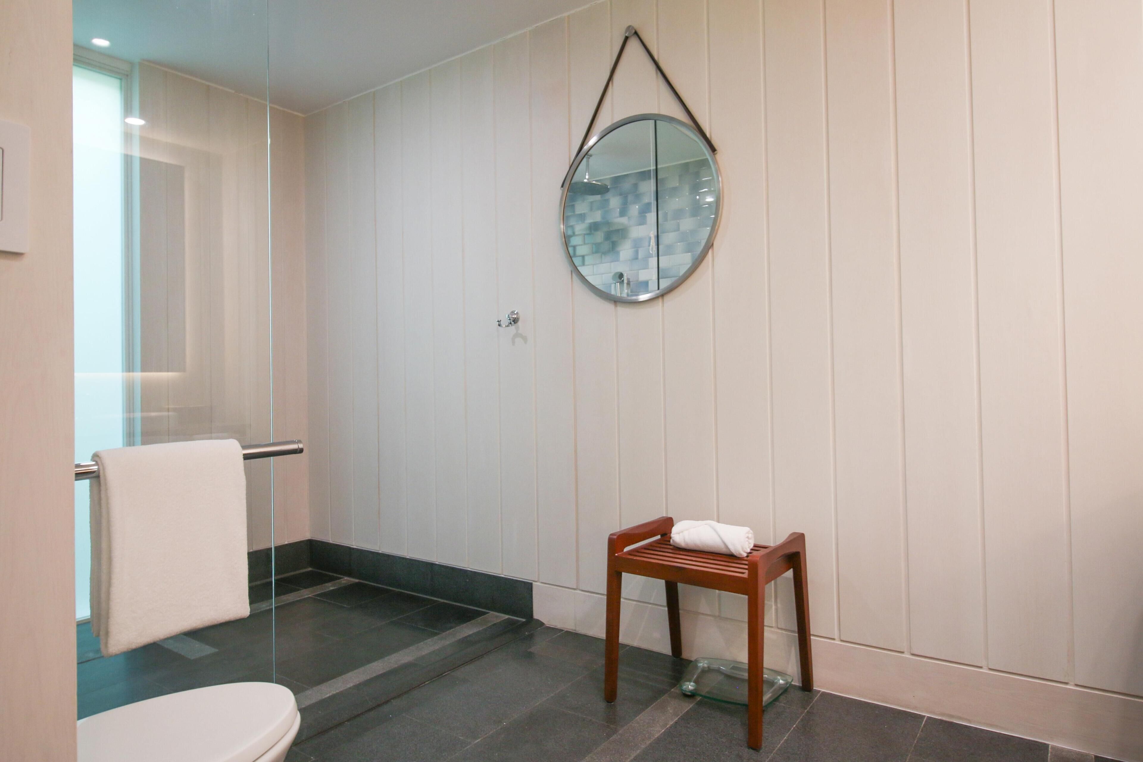 Ein Badezimmer mit Toilette, einer Duschkabine aus Glas, einem Holzstuhl und einem runden Spiegel an der Wand.