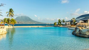 Ein Poolbereich mit klarem Wasser, ein Strand, Palmen und ein Berg im Hintergrund.