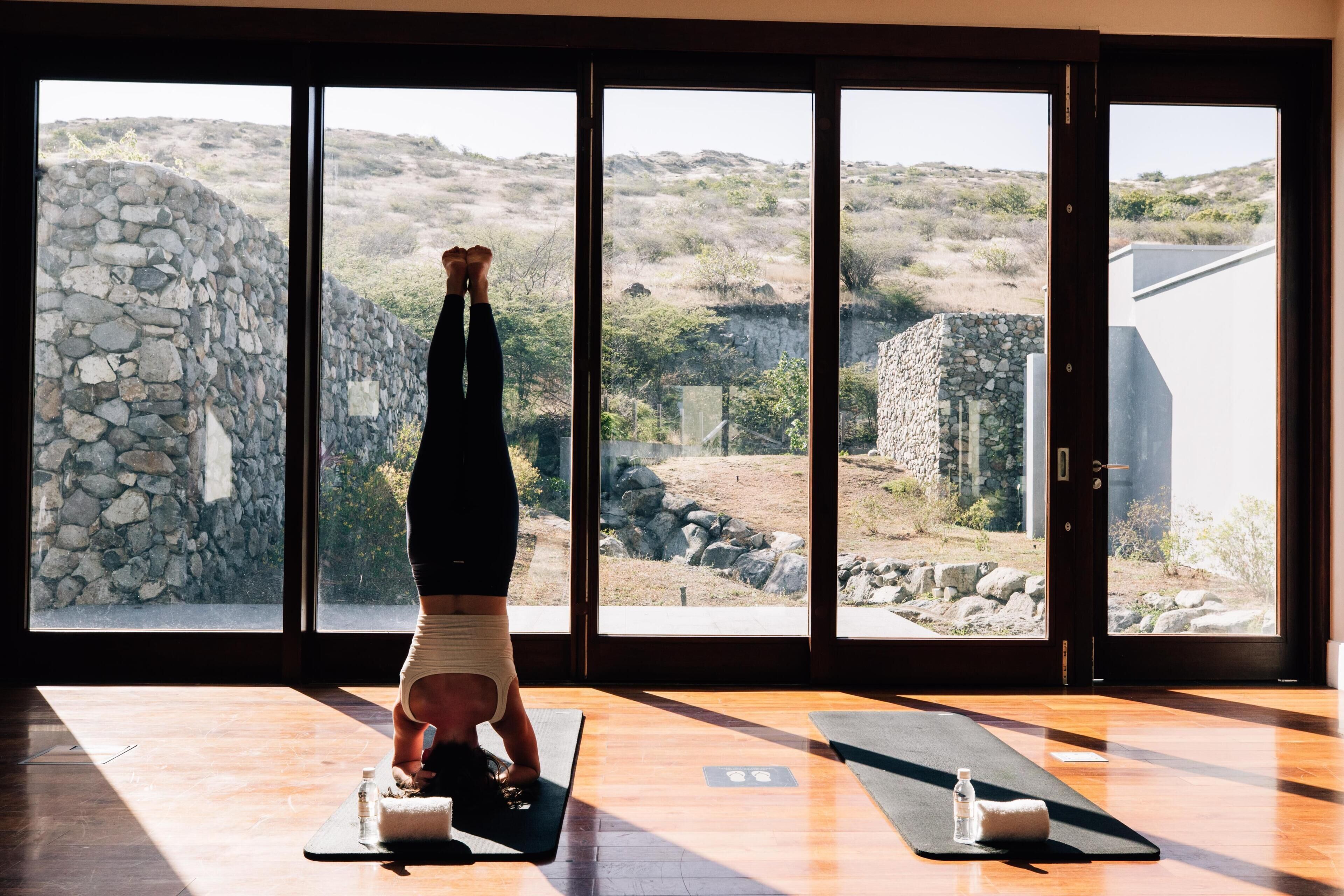Een persoon doet een handstand op een yogamat in een kamer met grote ramen en een stenen muur.