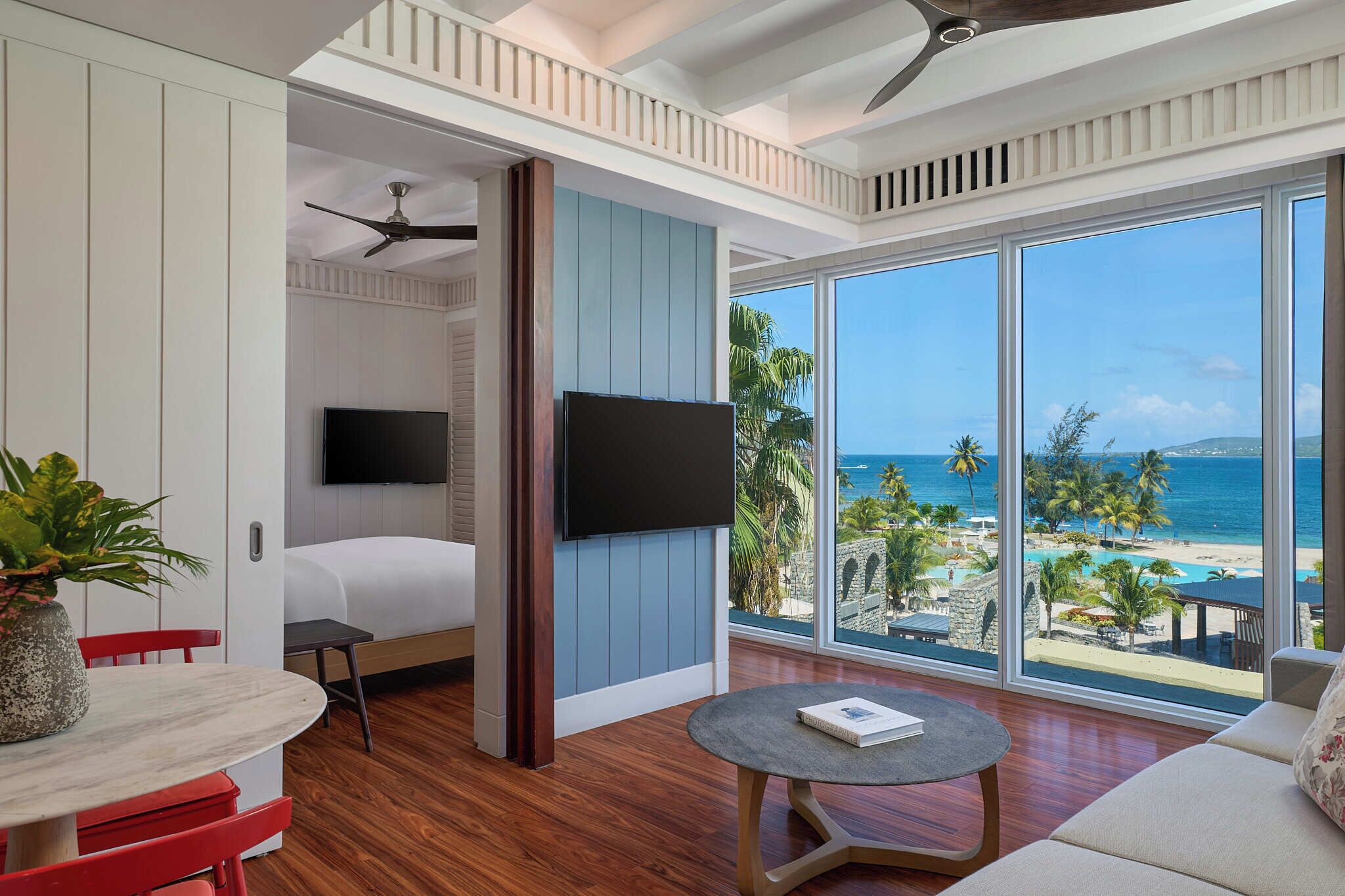 A hotel room with a bed, a TV, a dining table, a red chair, a round table, and a large window with a beach view.