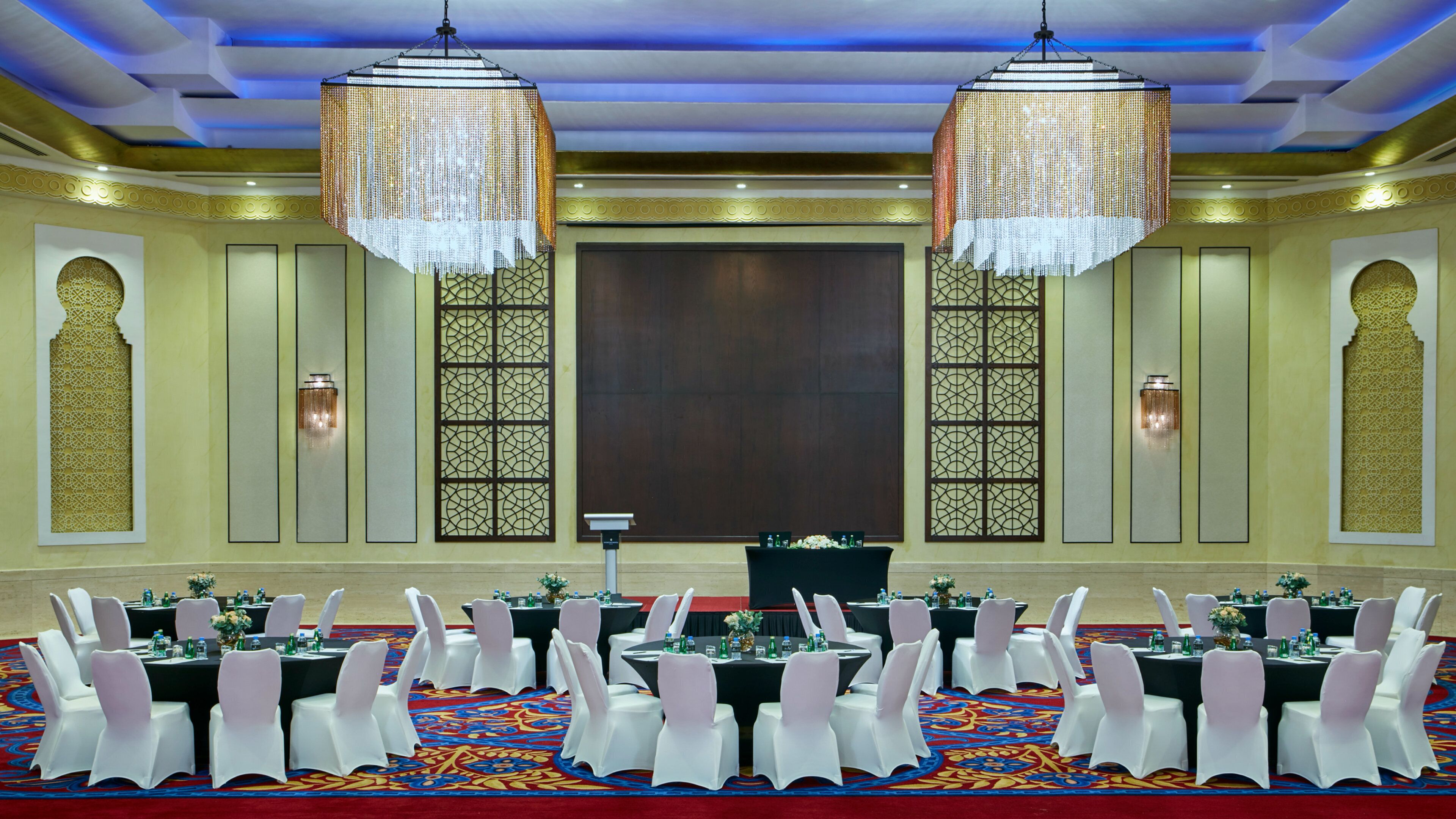 A banquet hall with round tables, white chairs, and chandeliers.