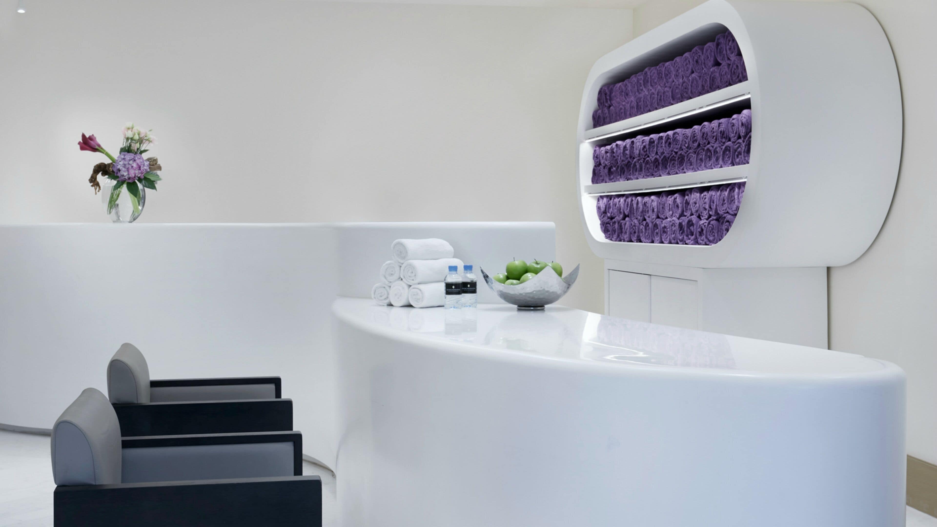 A reception desk with a white countertop, a vase with flowers, and a wall-mounted shelf with rolled towels.