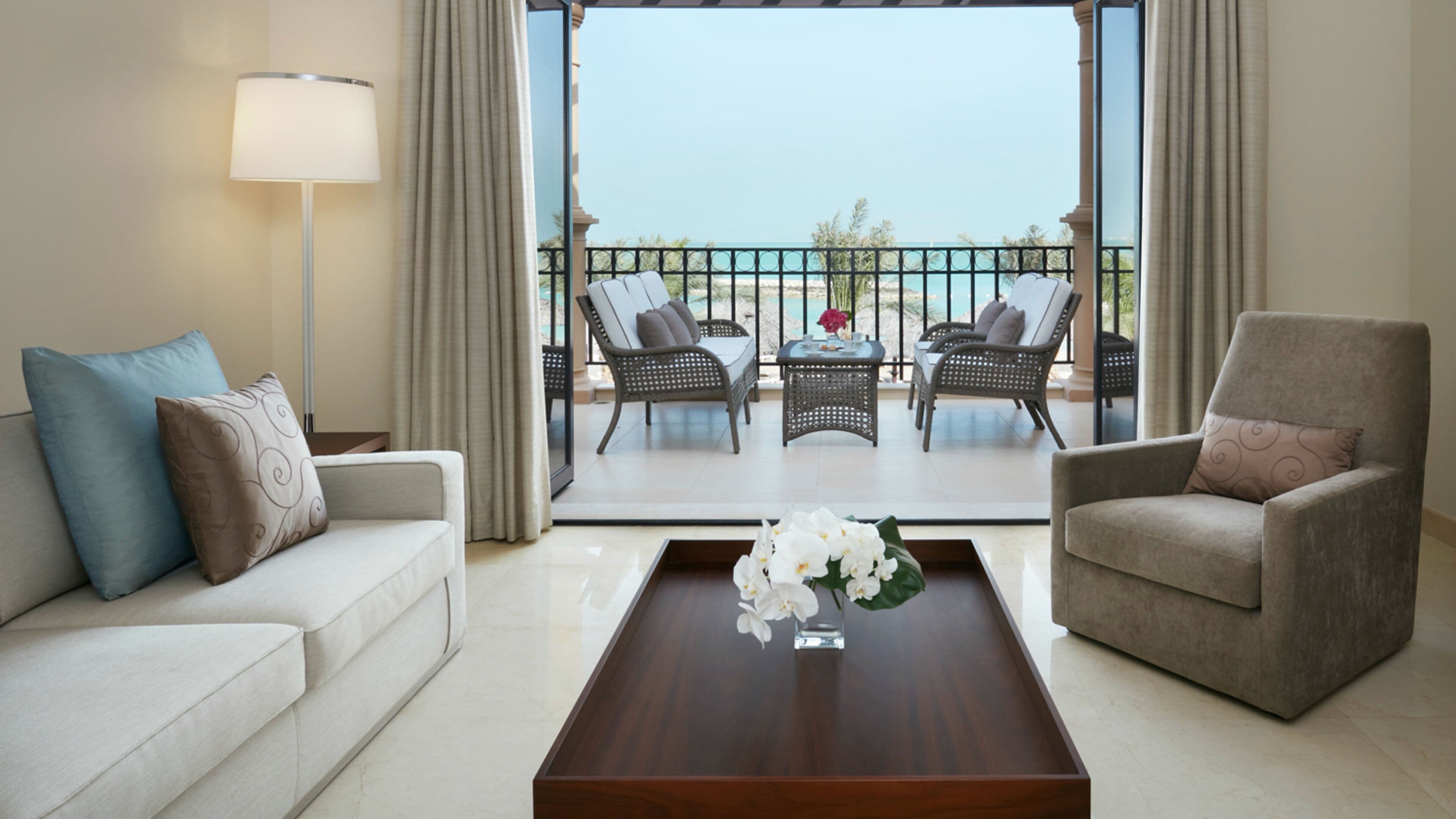 Une chambre d’hôtel avec un balcon, un canapé, un fauteuil et une table basse agrémentée de fleurs.