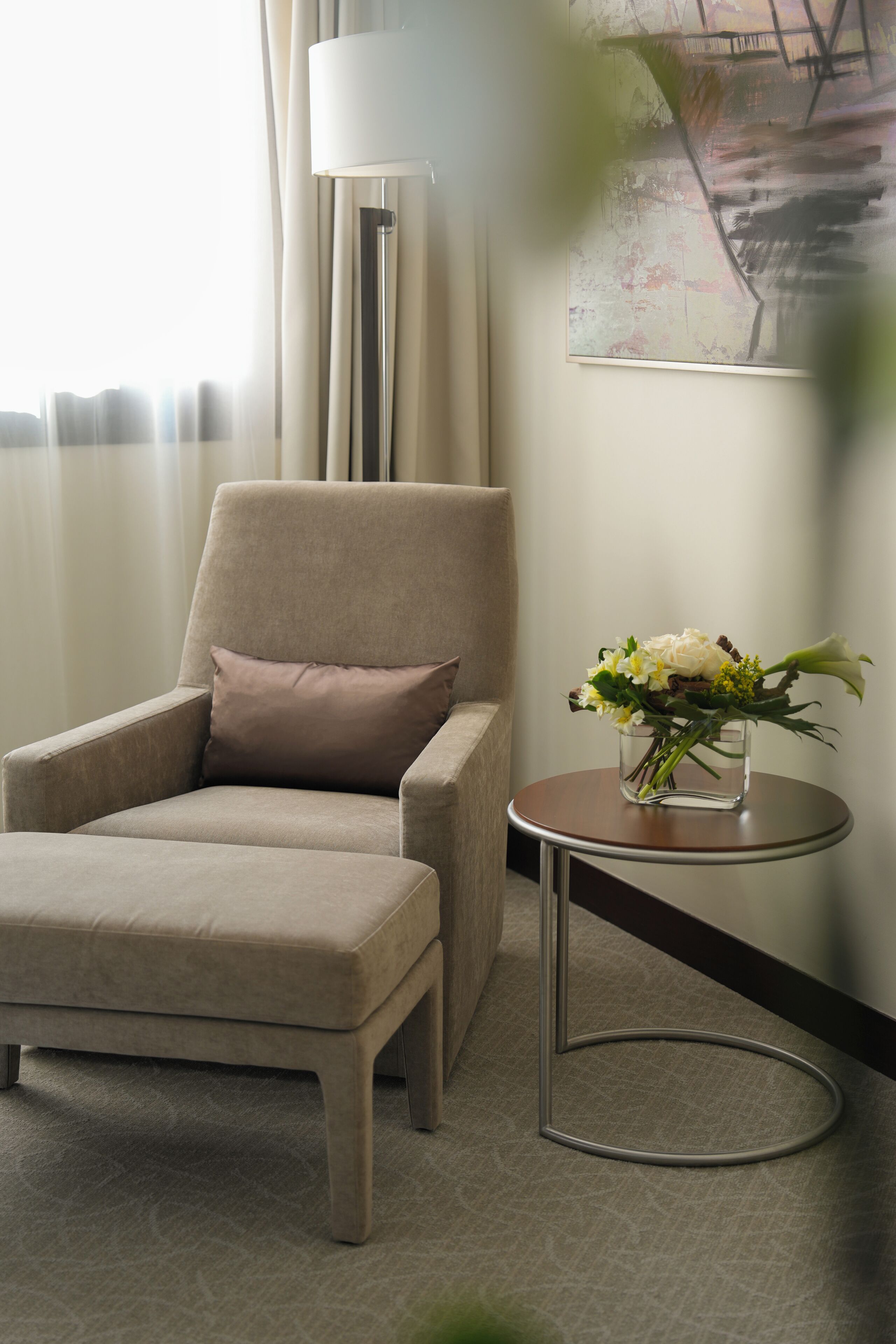 A hotel room with a beige armchair, a small round table with a vase of flowers, and a floor lamp.