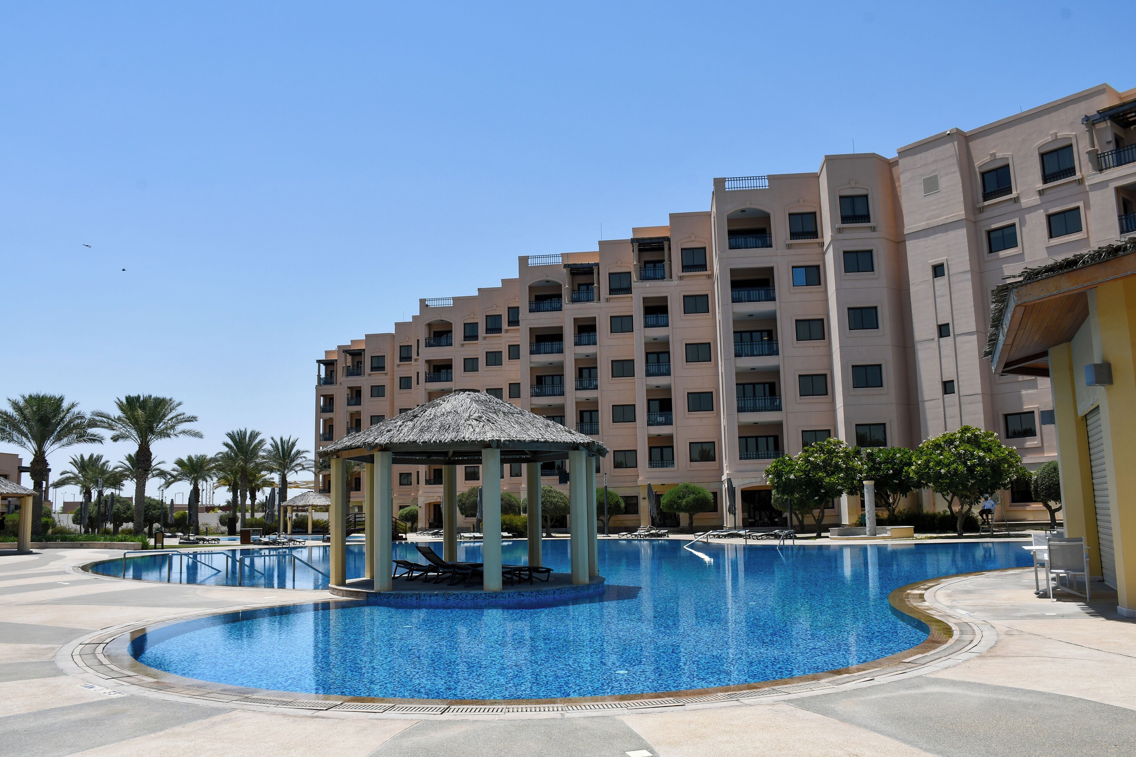A swimming pool with a gazebo, palm trees, and a multi-story hotel building in the background.