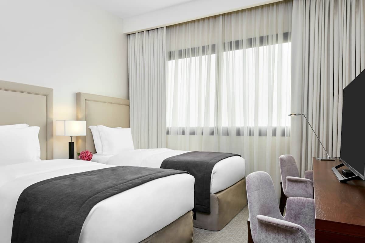 Une chambre d’hôtel avec deux lits, un bureau, une chaise et une grande fenêtre avec des rideaux légers.