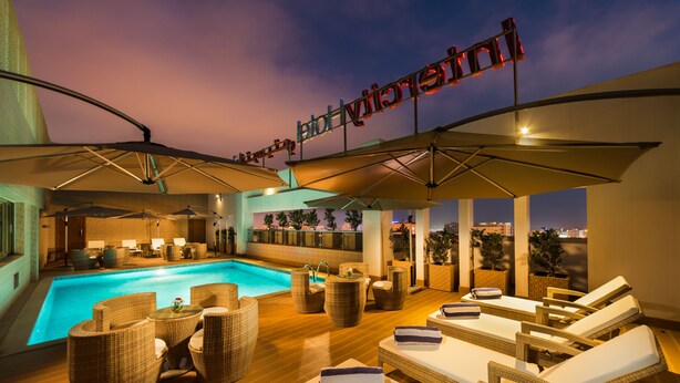 Un espace piscine sur le toit, avec des chaises longues et des parasols, un bar et une vue sur les toits de la ville au crépuscule.