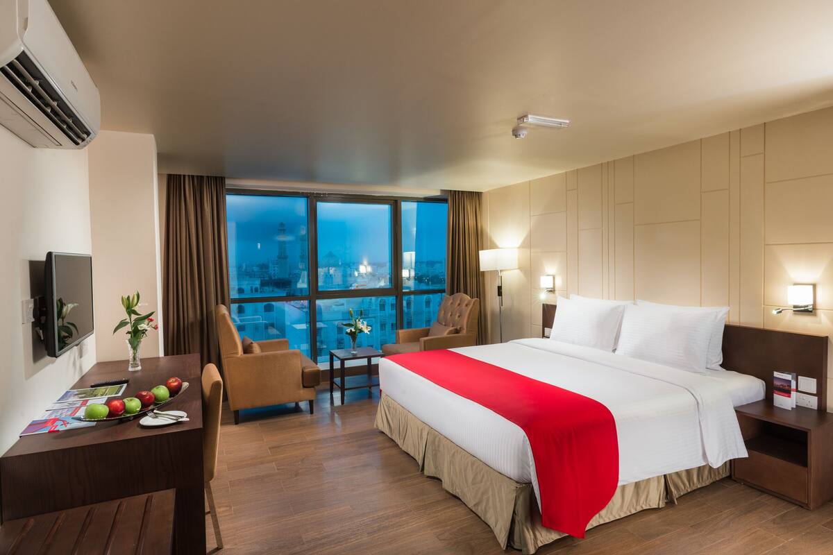 Een hotelkamer met een groot bed, een bureau met een bloemenvaas, een stoel, en een stadsgezicht door grote ramen.